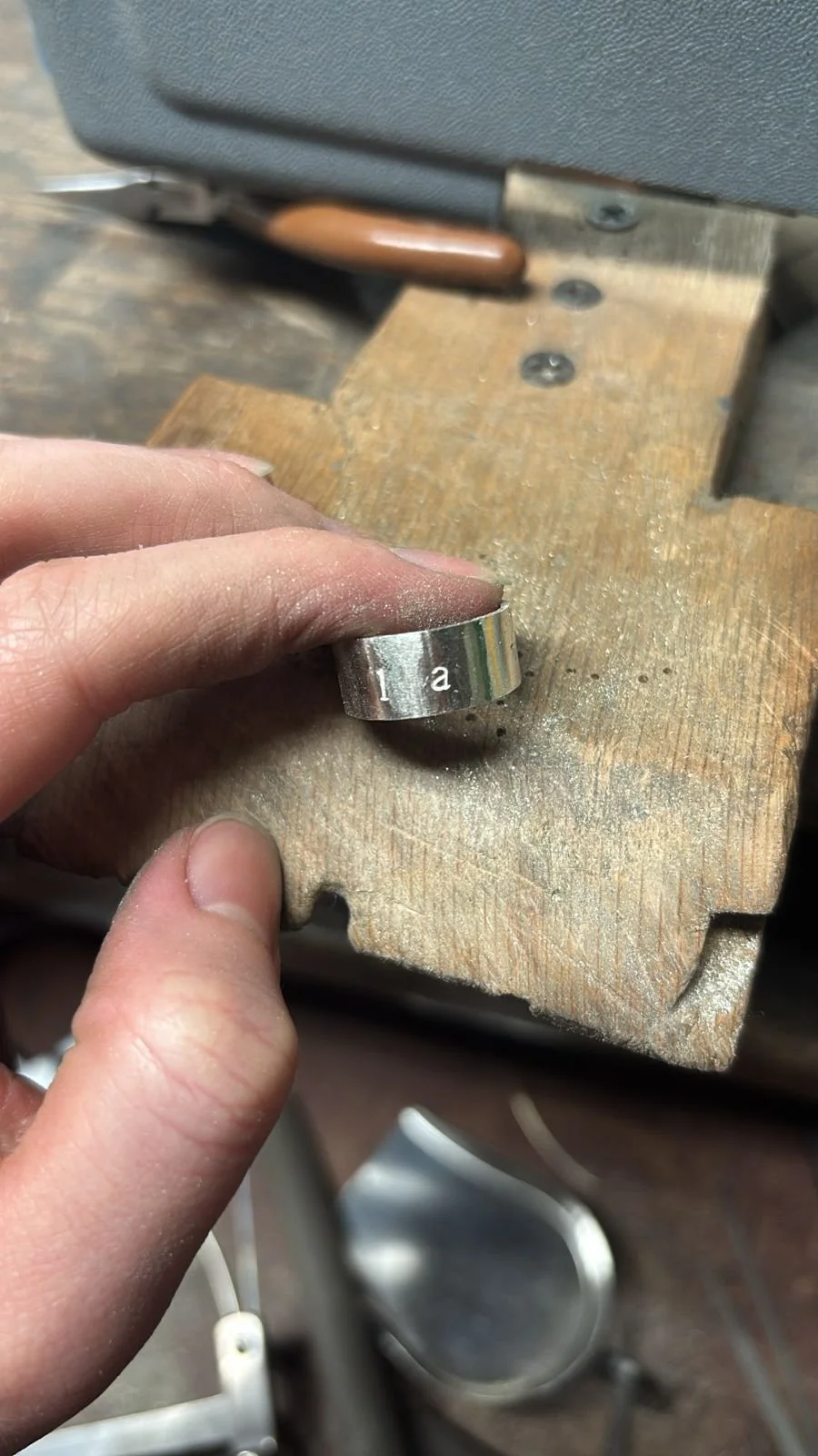 Persona sosteniendo una pieza metálica que tiene letras grabadas, en un taller con banco de trabajo de madera y herramientas en el fondo.