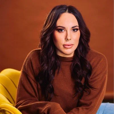 A young woman with long dark wavy hair wearing a brown sweater sitting on a yellow chair with a warm brown background.