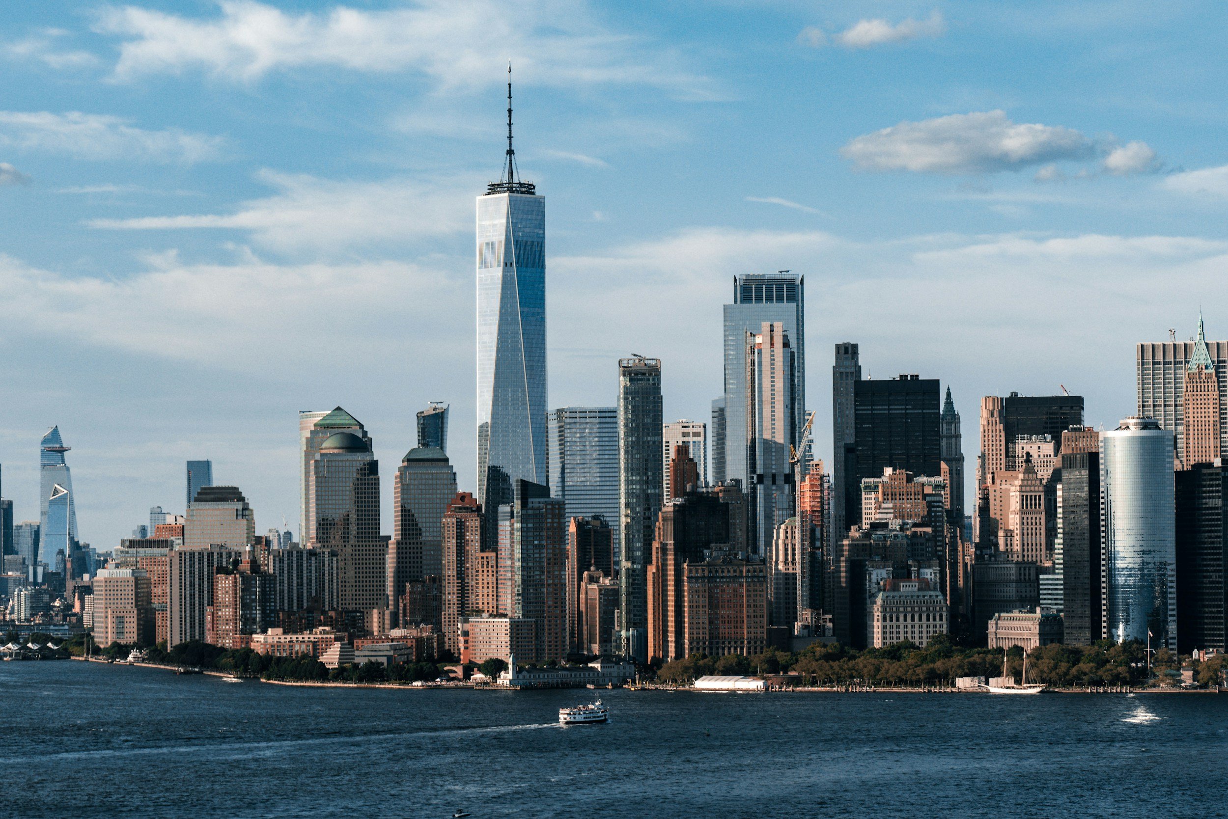 Skyline of Manhattan with One World Trade Center in the center, across a body of water, with several boats, under a partly cloudy sky.