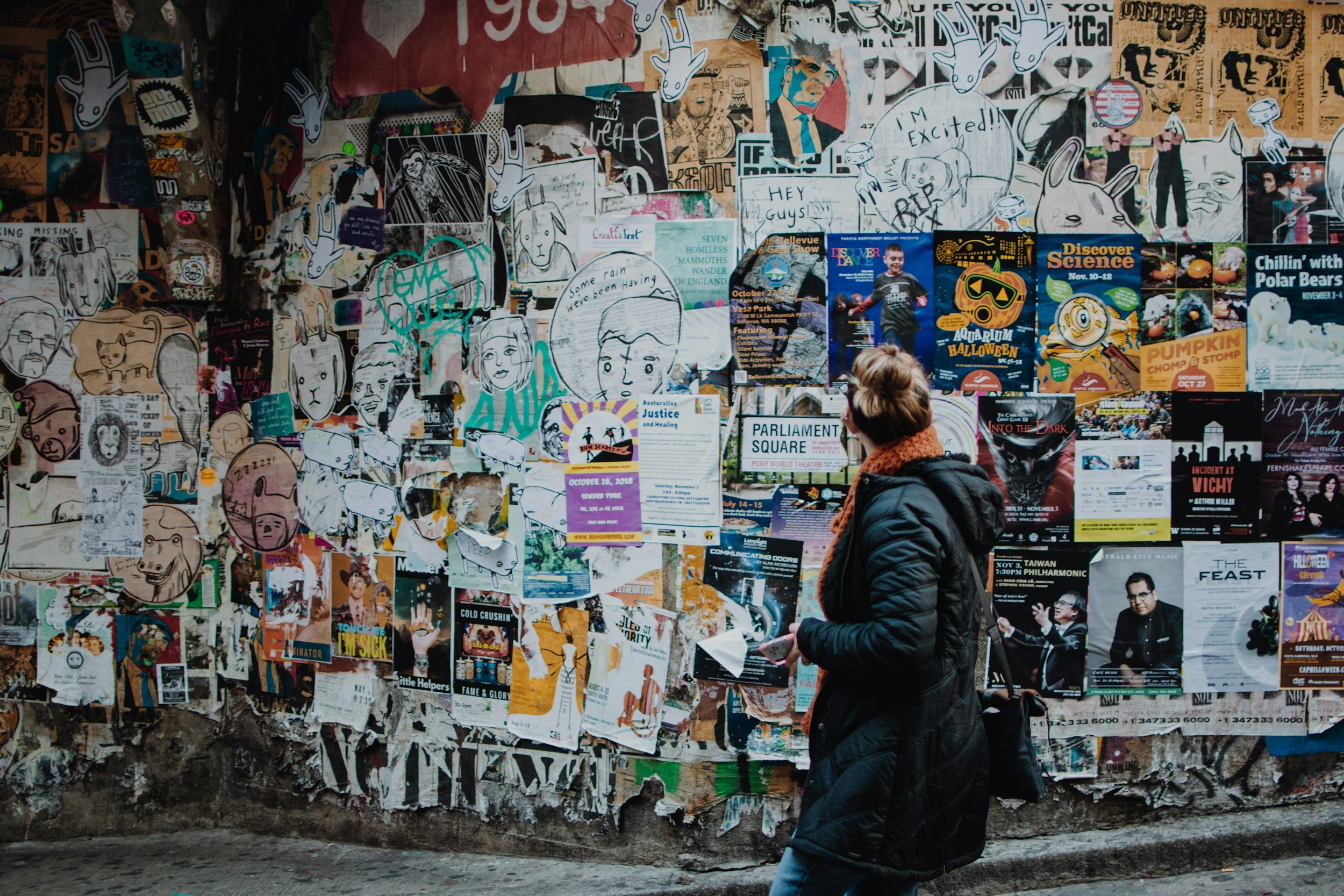 A woman with short hair wearing a black quilted coat and orange scarf, standing in front of a colorful wall covered with posters, drawings, and flyers, reading or viewing the wall.