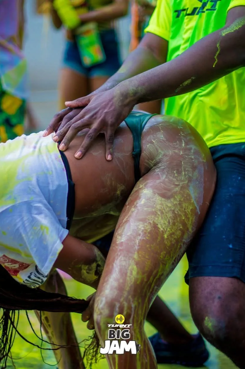 A person playing a messy game, covered in yellow powder, leaning forward while another person is giving a massage or hug, both participating in the event at a colorful outdoor gathering with the event 'TURANT BIG JAM' logo at the bottom.
