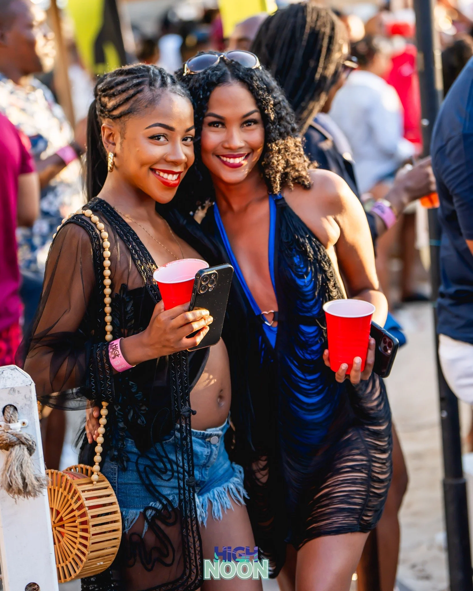 Two young women smiling and holding red cups at an outdoor event, with other people in the background.