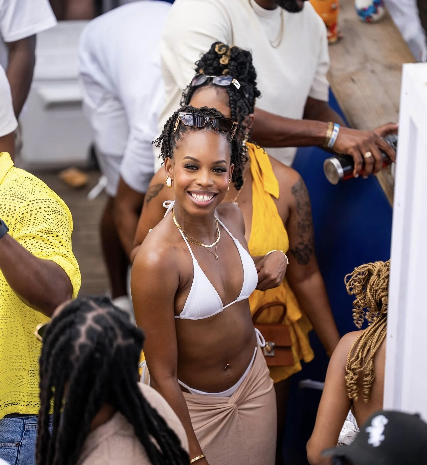 A smiling woman in a white bikini top and beige skirt at a social gathering, with people around her.