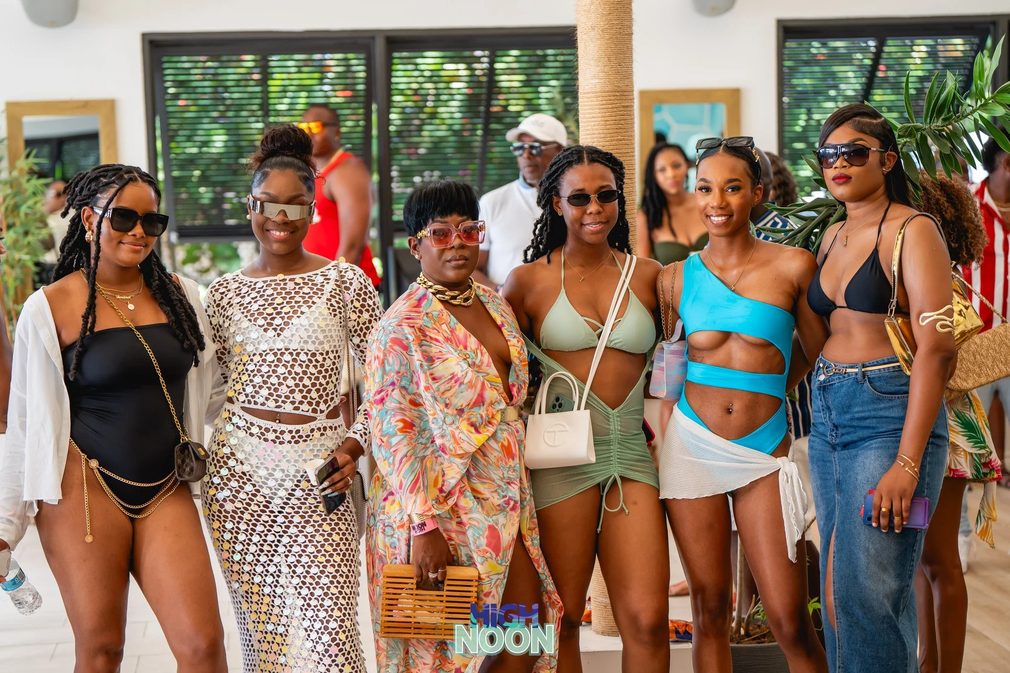 Group of women in swimwear and summer outfits posing indoors with plants and people in the background.