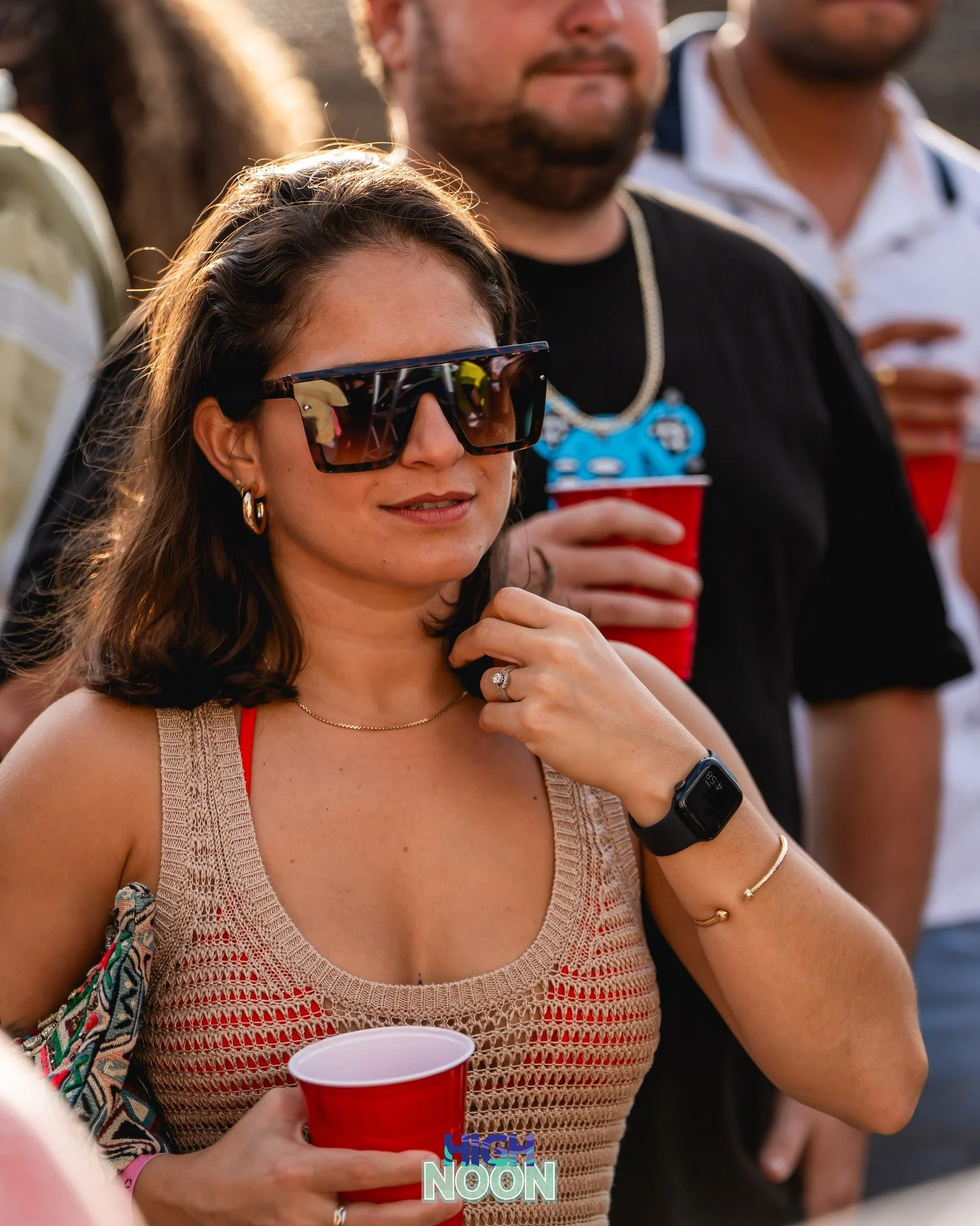 Woman wearing large sunglasses, a beige crochet top, and jewelry, holding a red cup at an outdoor event, with others in the background.