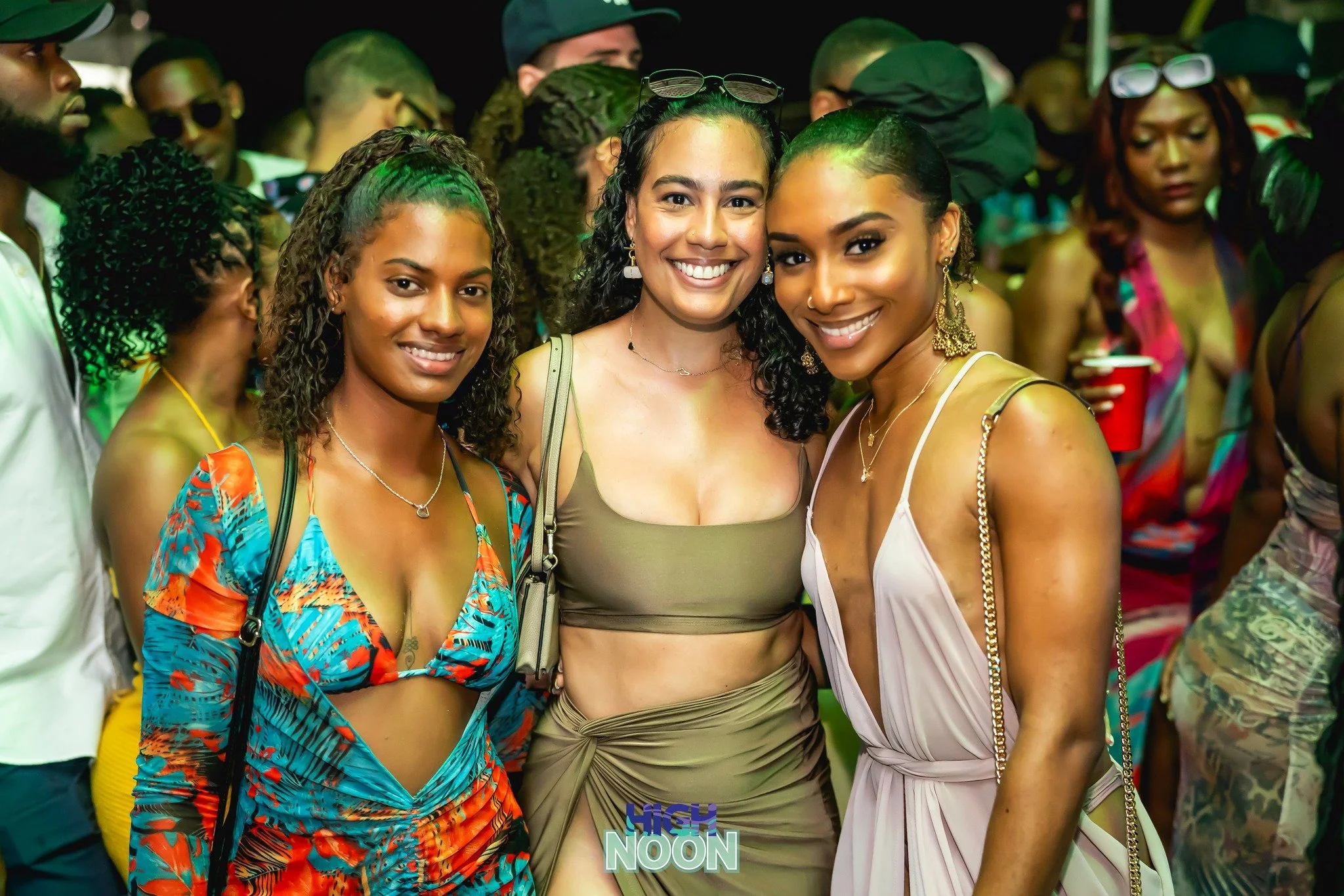 Three women smiling at a lively outdoor event, with people in colorful outfits and accessories in the background.