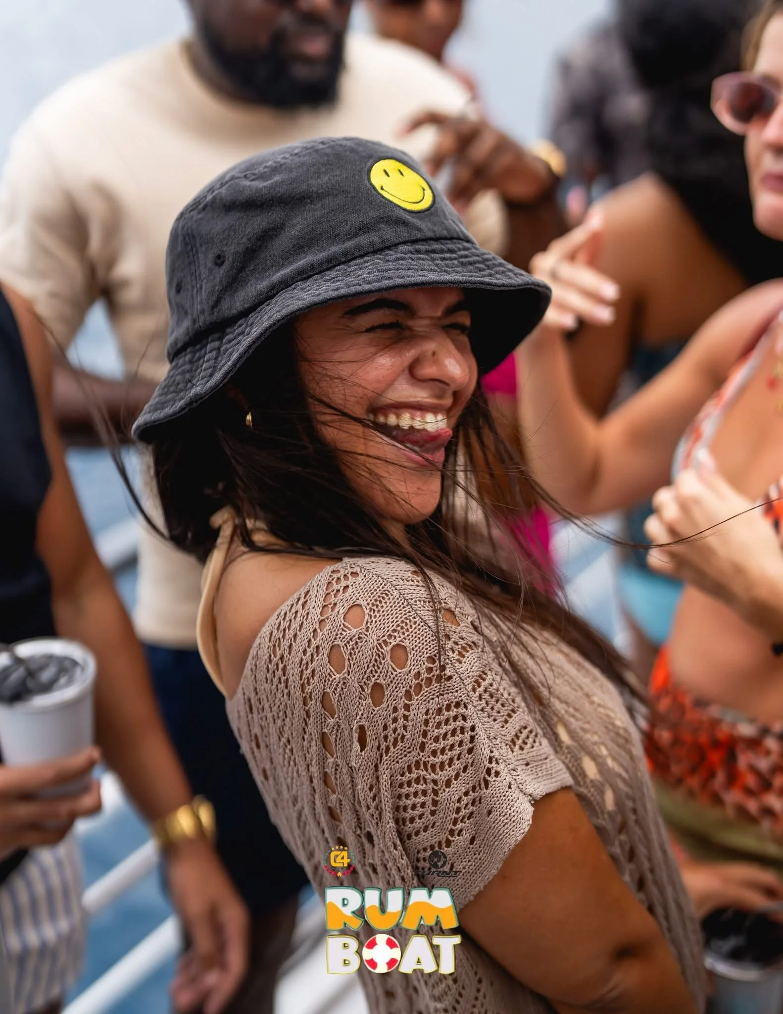 A woman with long dark hair wearing a black bucket hat with a yellow smiley face, smiling and laughing among a group of people on a boat.