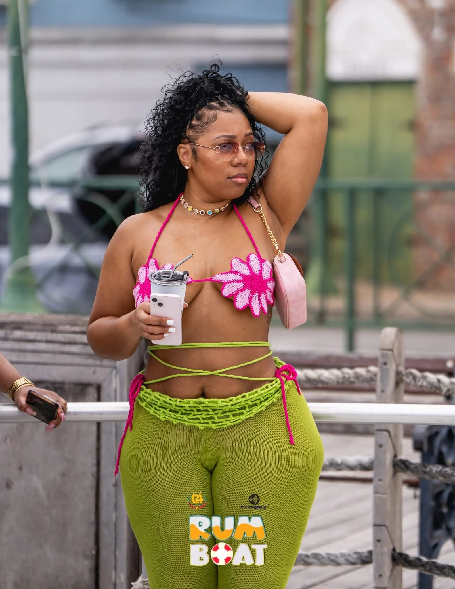 A woman with curly hair wearing sunglasses, a pink floral bikini top, green pants, and accessories, standing outdoors holding a drink, with a background of a railing and blurred outdoor scene.