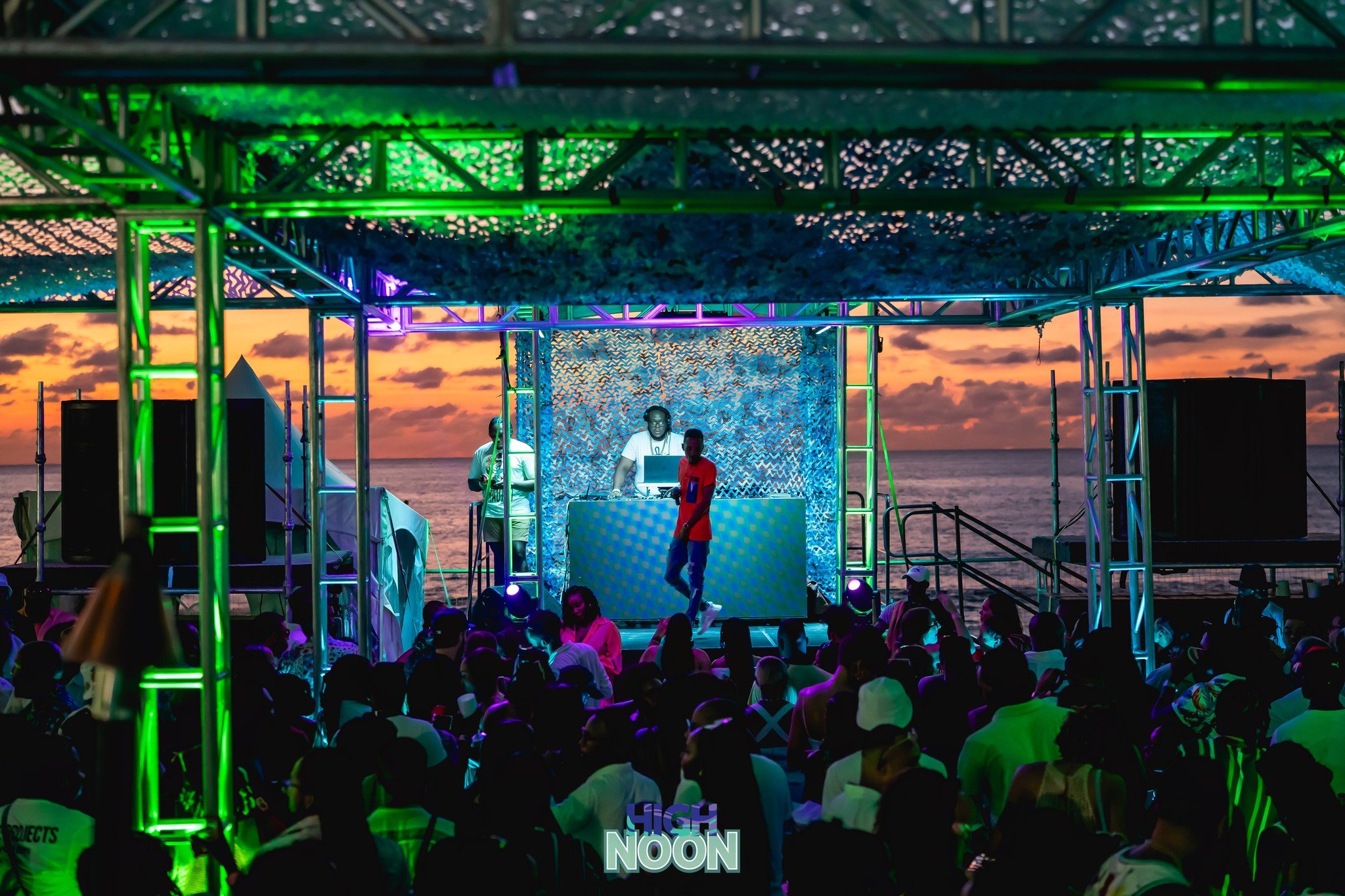 A sunset party scene on the beach with a DJ playing music behind a colorful, illuminated stage, and a crowd of people dancing and enjoying the event, with the ocean and sky in the background.