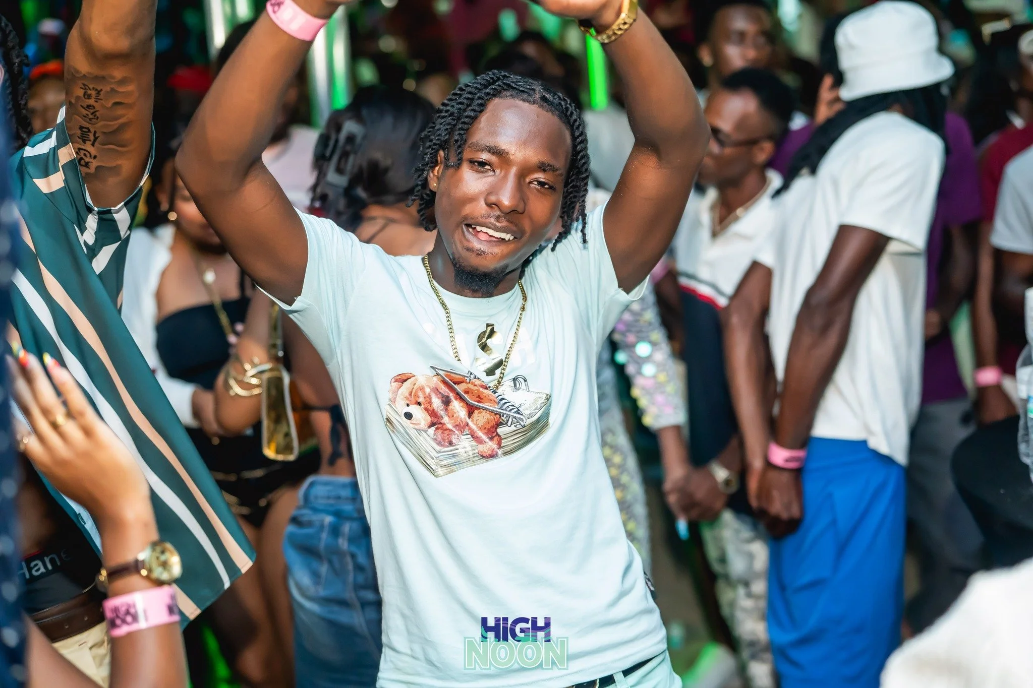 A young man with braided hair dancing at a crowded event, wearing a white t-shirt with a stuffed animal print, gold chains, and a gold watch, surrounded by other people in casual attire.