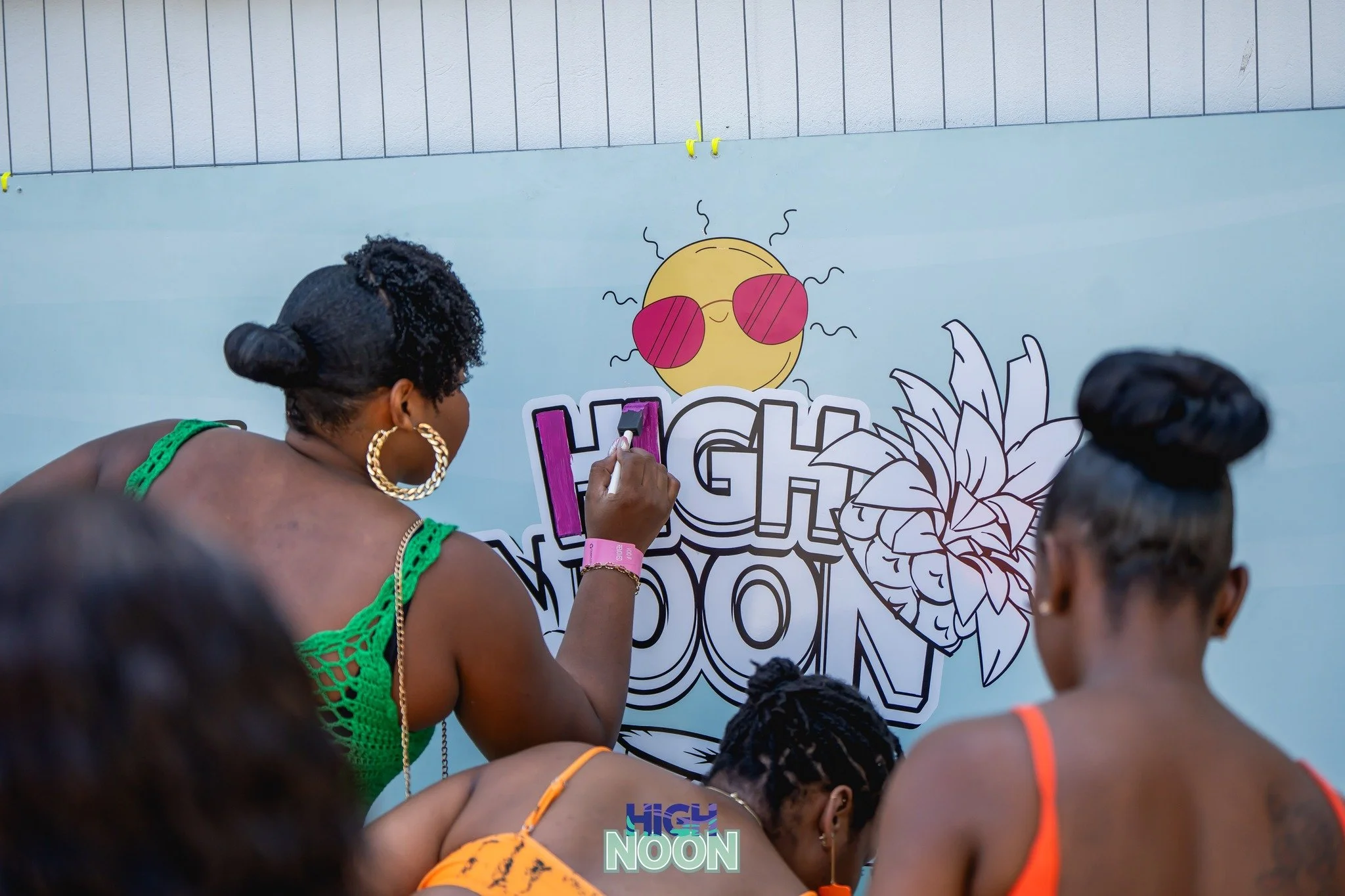 Women in colorful outfits painting a mural that says 'High Noon' with a smiling sun wearing sunglasses and a large flower.