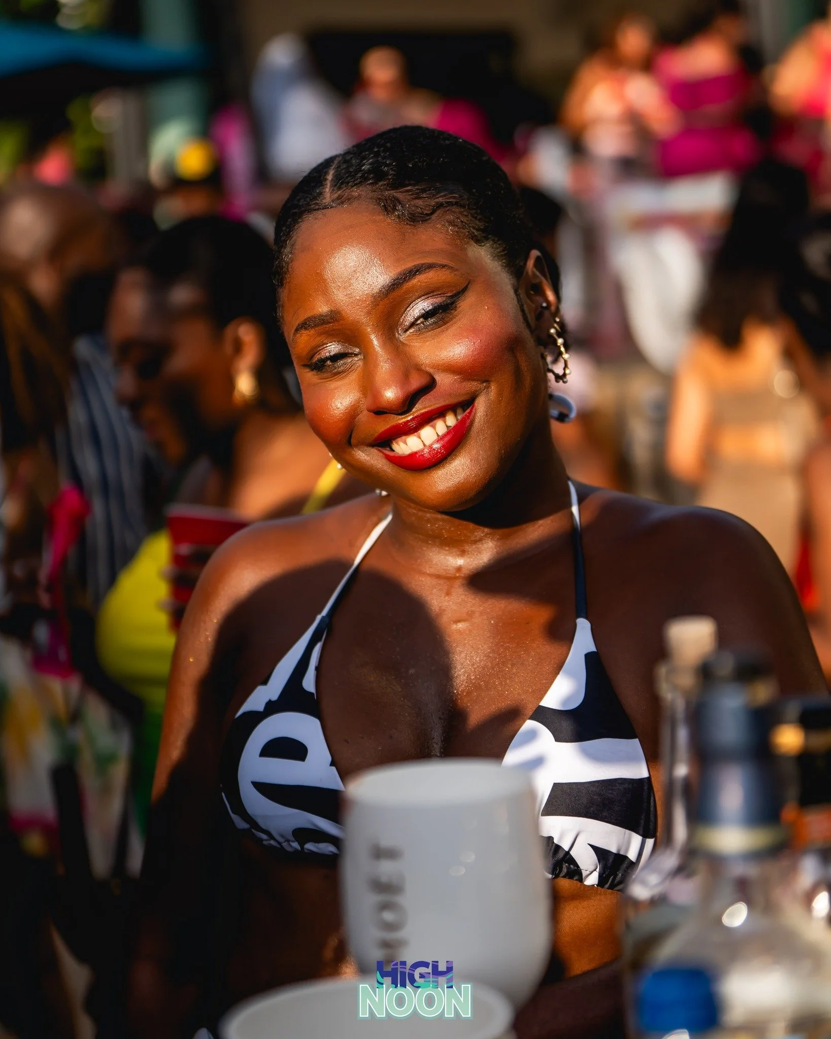 A woman smiling with lipstick at an outdoor gathering, surrounded by others in colorful clothing.