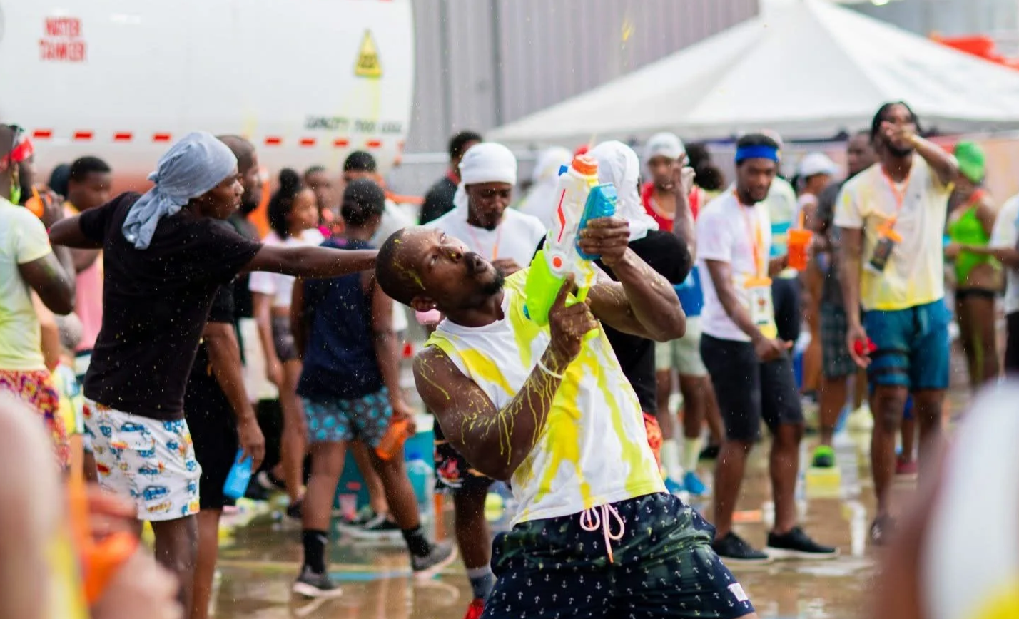 People participating in a water fight, with a man in the foreground holding a water gun and splattered with water and yellow paint, while others in the background cheer and spray water.