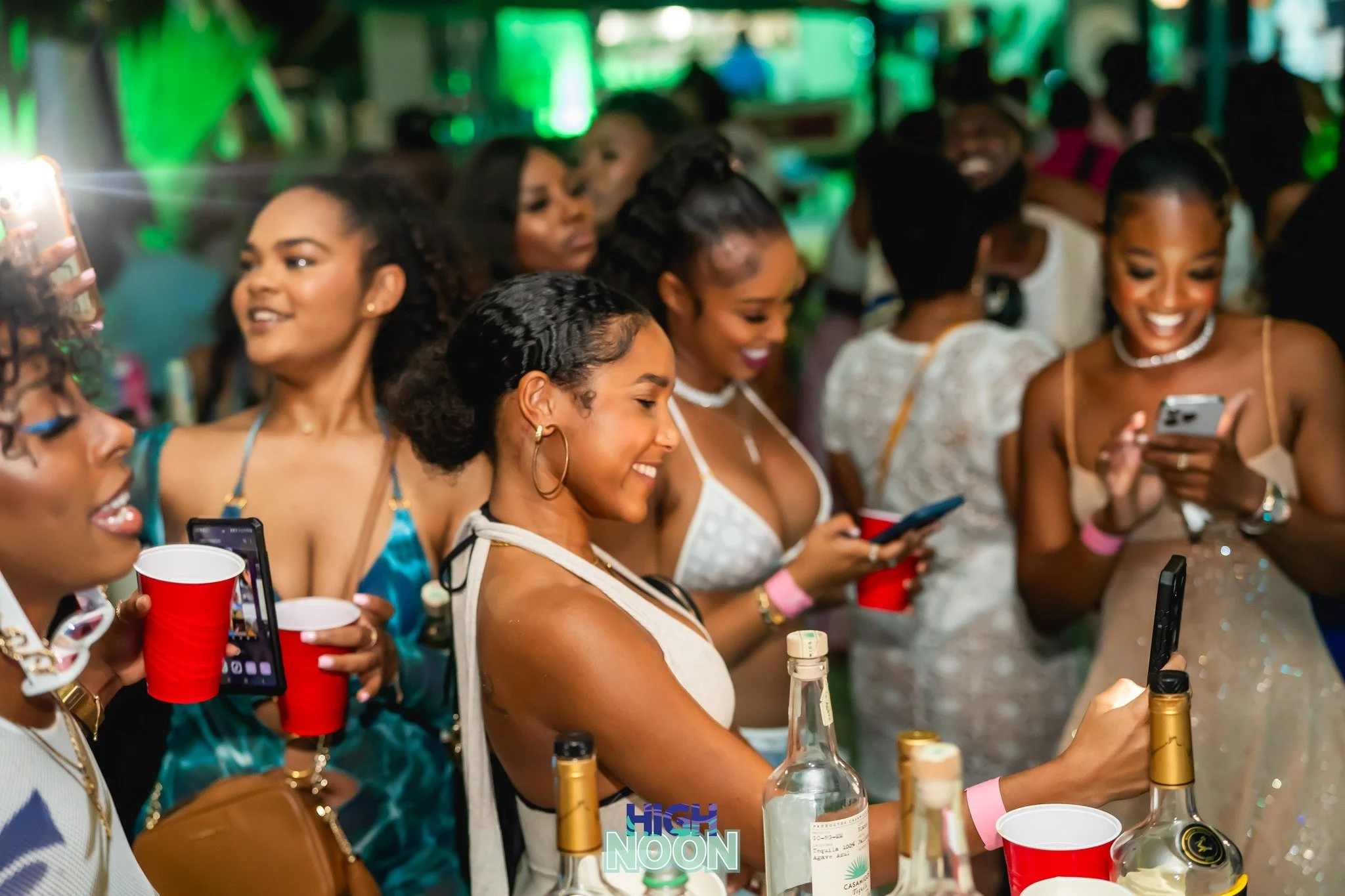 Group of women at a party, enjoying drinks, smiling, and taking photos with their phones.