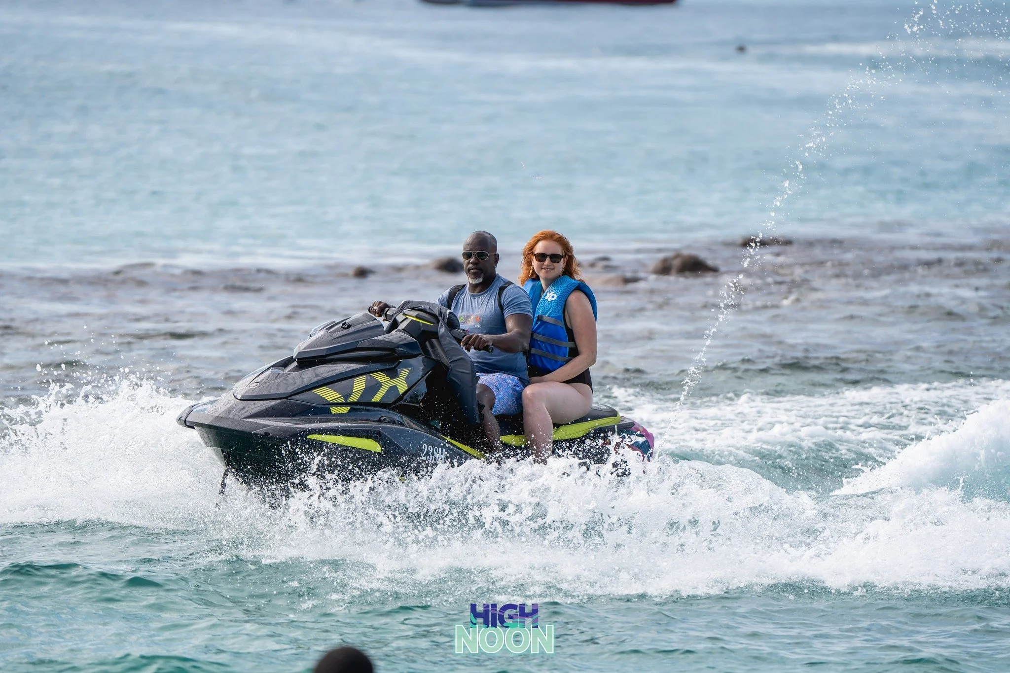 A man and woman riding a jet ski on the water, both wearing life jackets, with the woman smiling and sunglasses.