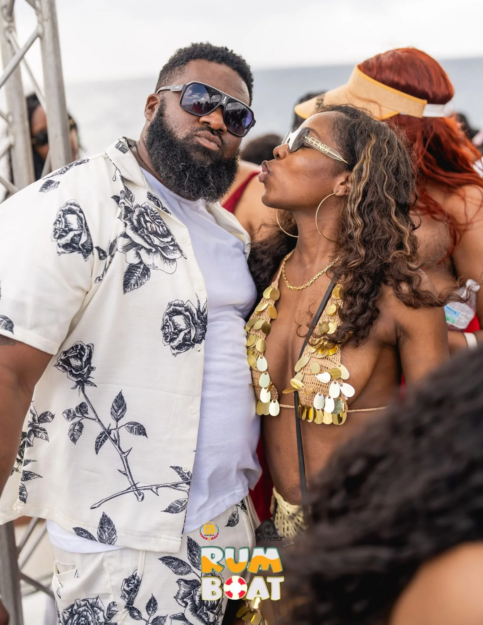 A man and woman standing close together at a beach event, with the man wearing sunglasses and a white floral shirt, and the woman wearing sunglasses and a gold sequin top, both engaging in a playful moment.