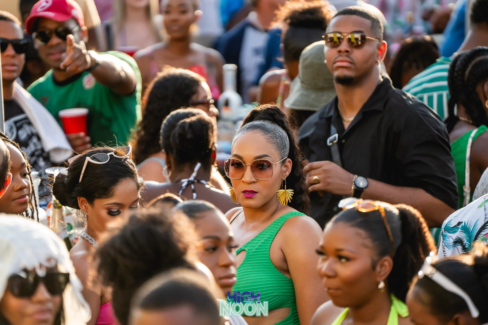 A large crowd of people at an outdoor event, many wearing colorful clothing, sunglasses, and accessories, with some holding drinks. The focus is on a woman in a green dress with large sunglasses and gold earrings, surrounded by others enjoying the ev