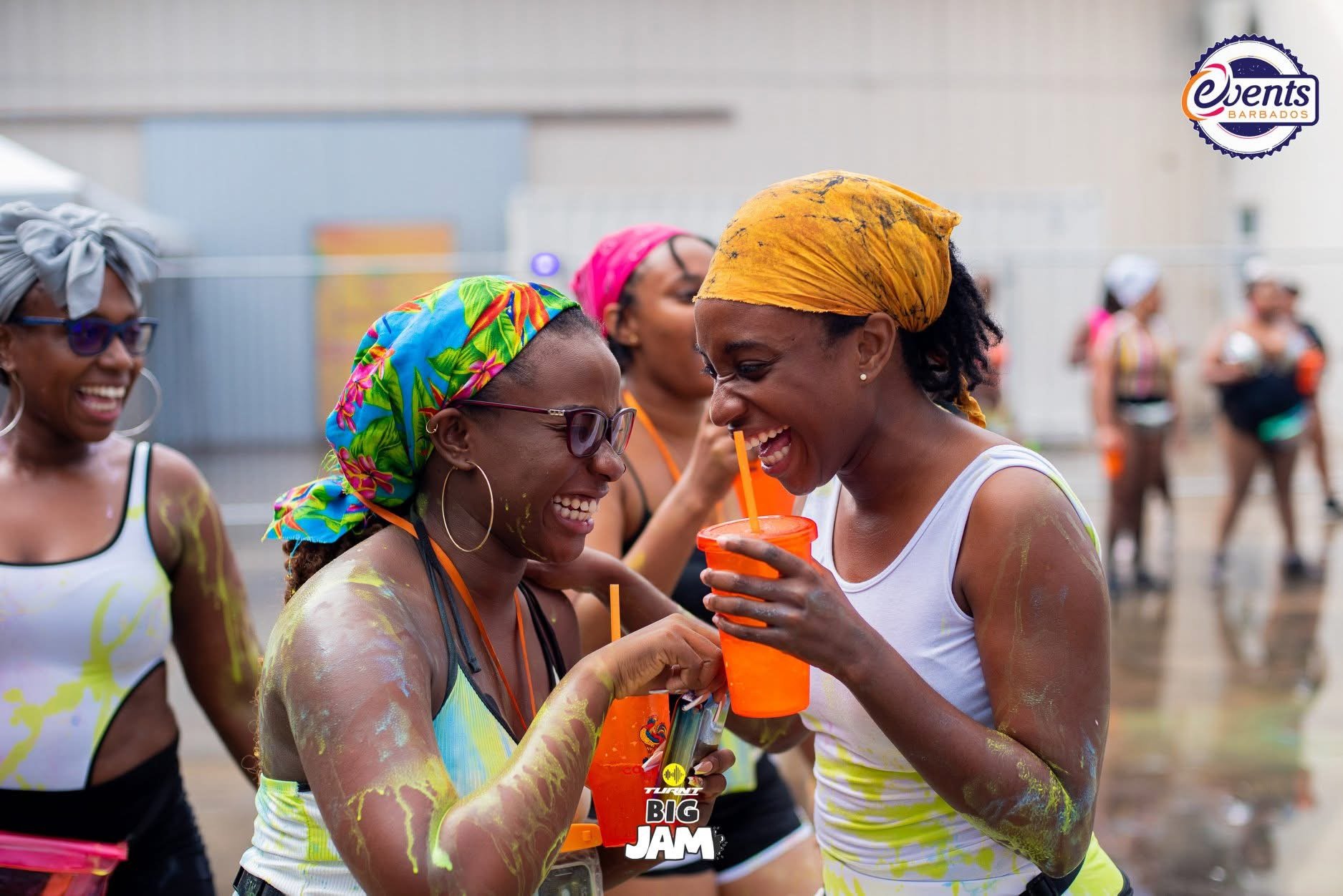 Women celebrating at an outdoor event with colorful headscarves and drinks, covered in yellow powder, with a logo for 'Event Barbados' and 'Big Jam' in the corner.