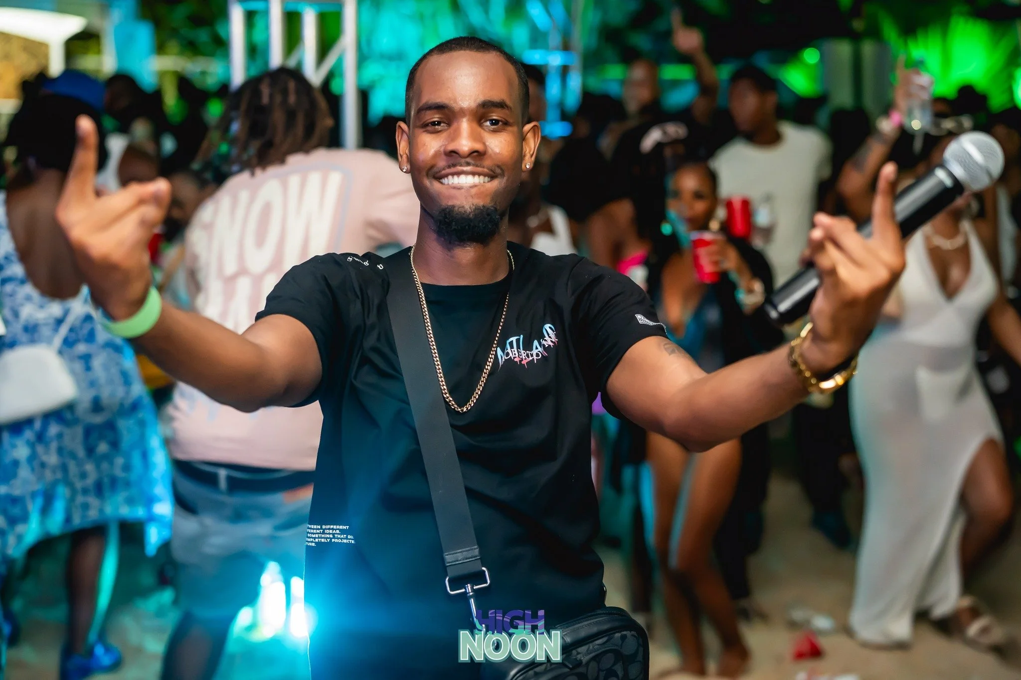 A smiling man holding a microphone at a lively party with people dancing and socializing in the background.