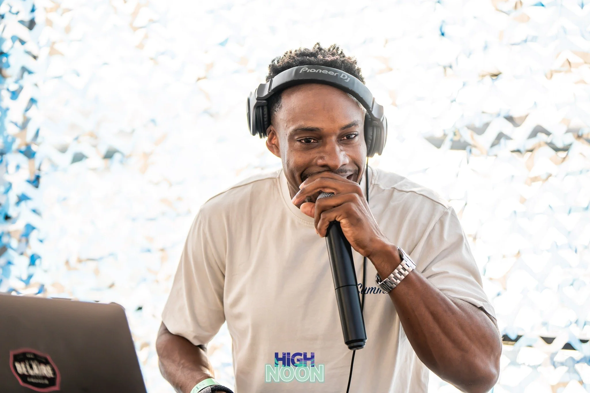 A man with dark curly hair wearing a Pioneer DJ headset, holding a microphone, with a laptop in front of him and a patterned background.