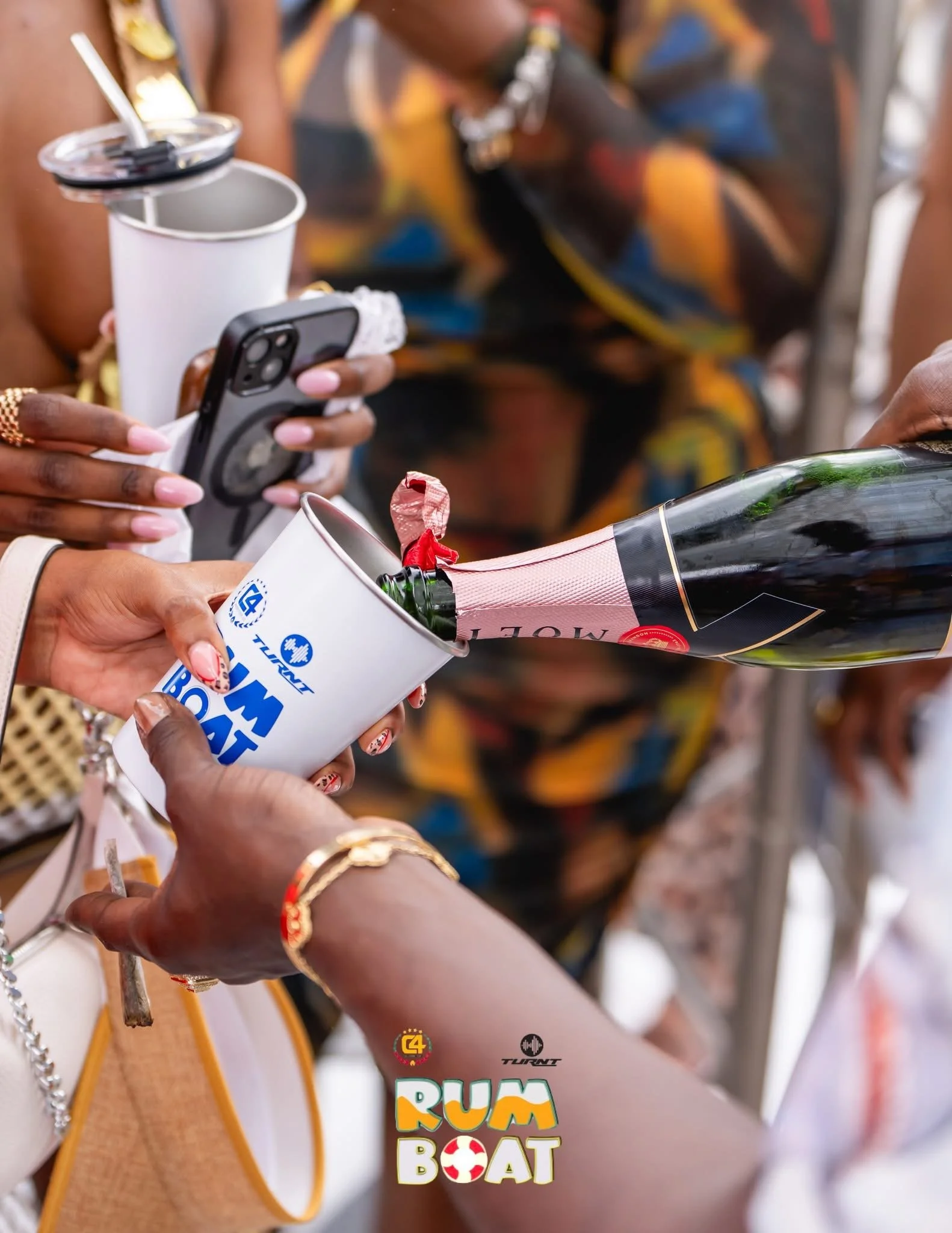 People celebrating, with one person pouring champagne into a paper cup held by another person, in front of a colorful skirt, with a logo that reads 'RUM BOAT' at the bottom.