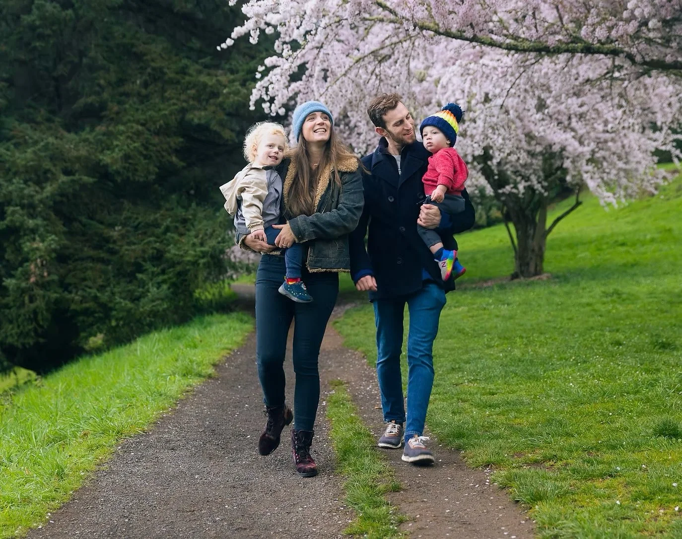 A family of four walking outdoors on a dirt path near a flowering tree, with green grass and trees in the background.