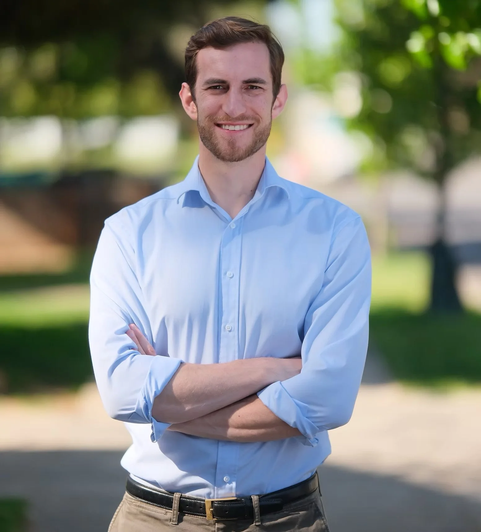 A smiling man with light brown hair and a beard, wearing a light blue dress shirt and khaki pants, standing outdoors with arms crossed in a park with green trees in the background.