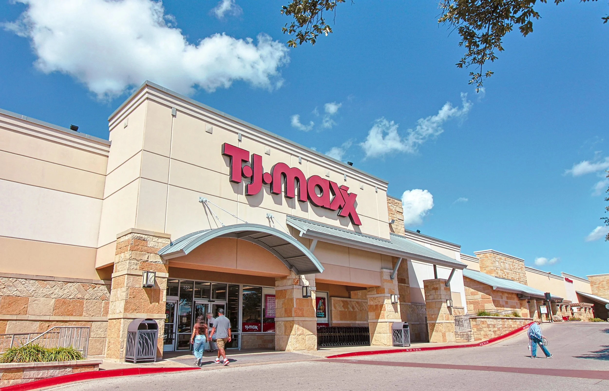 Exterior view of a T.J. Maxx store at Wolf Ranch Town Center in Georgetown, TX, with the store's sign prominently displayed, people entering and walking outside on a sunny day with a clear blue sky.