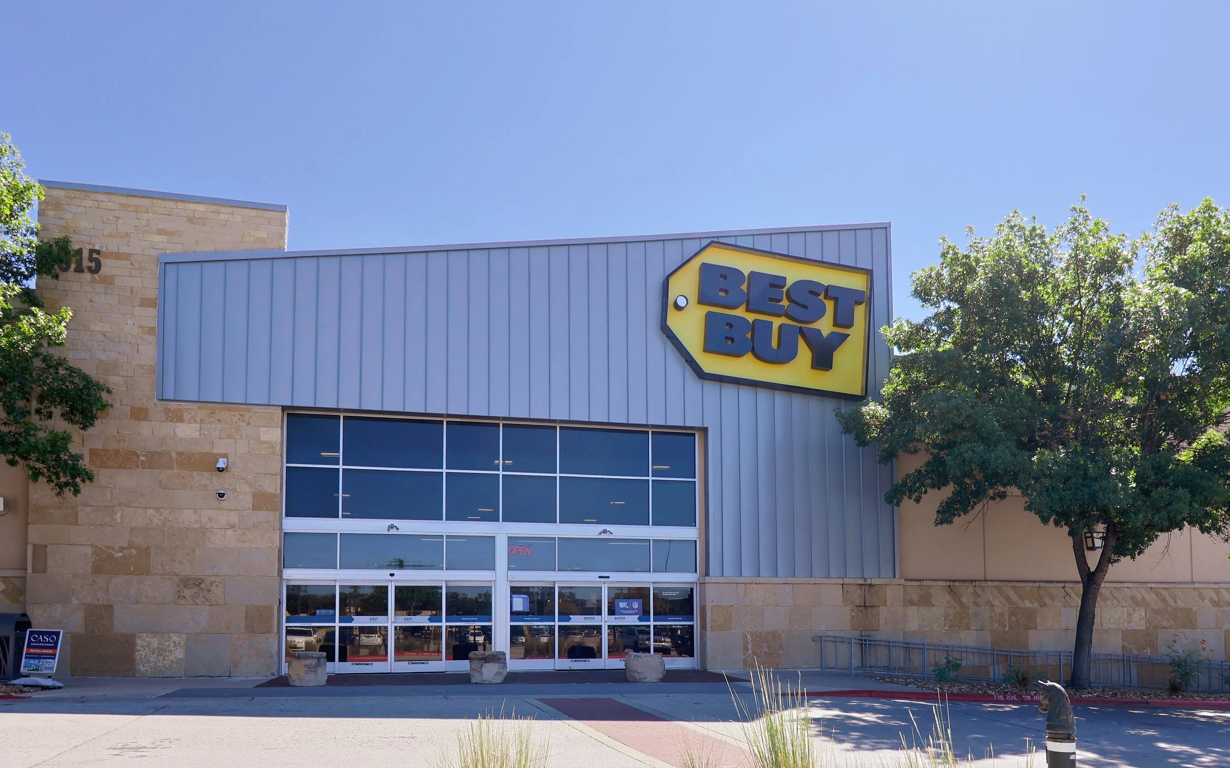 Exterior view of a Best Buy electronics retail store  at Wolf Ranch Town Center in Georgetown, TX, with a large sign, glass doors, and surrounding trees on a sunny day.