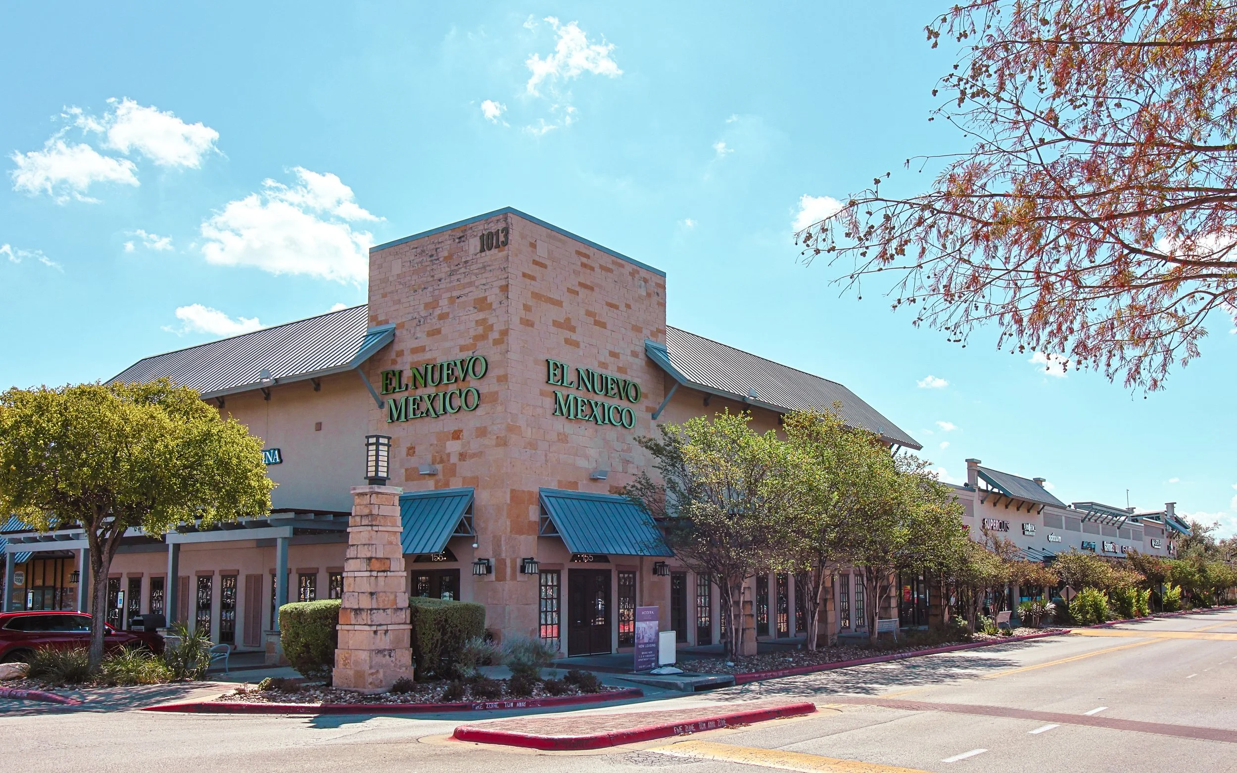 Exterior view of a shopping plaza with a sign that says "El Nuevo Mexico" at Wolf Ranch Town Center in Georgetown, TX, with shops and trees lining the street on a sunny day.