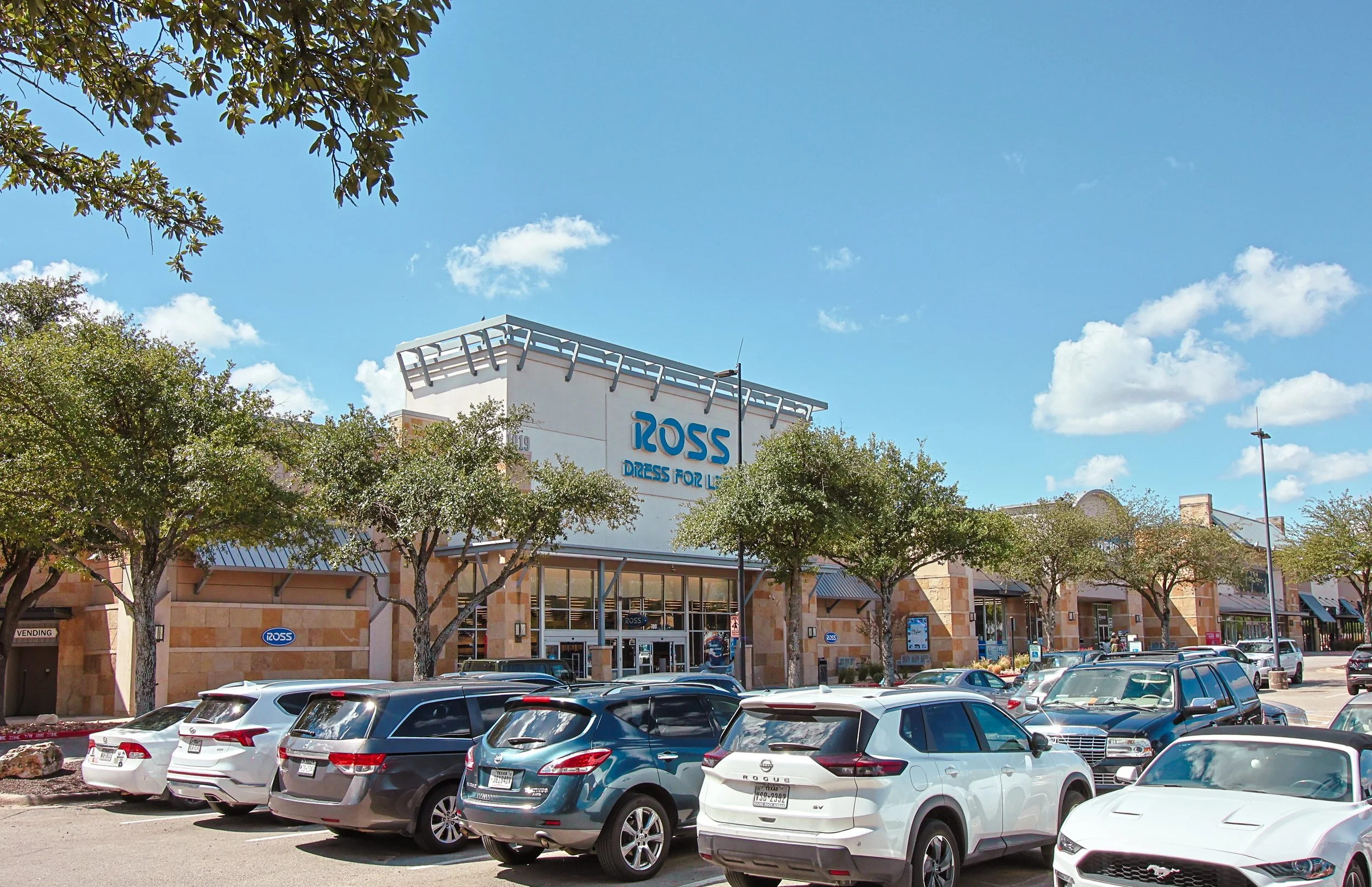 A shopping mall parking lot at Wolf Ranch Town Center in Georgetown, TX, with several parked cars in front of a Ross Dress for Less store, with a blue sky and some trees.