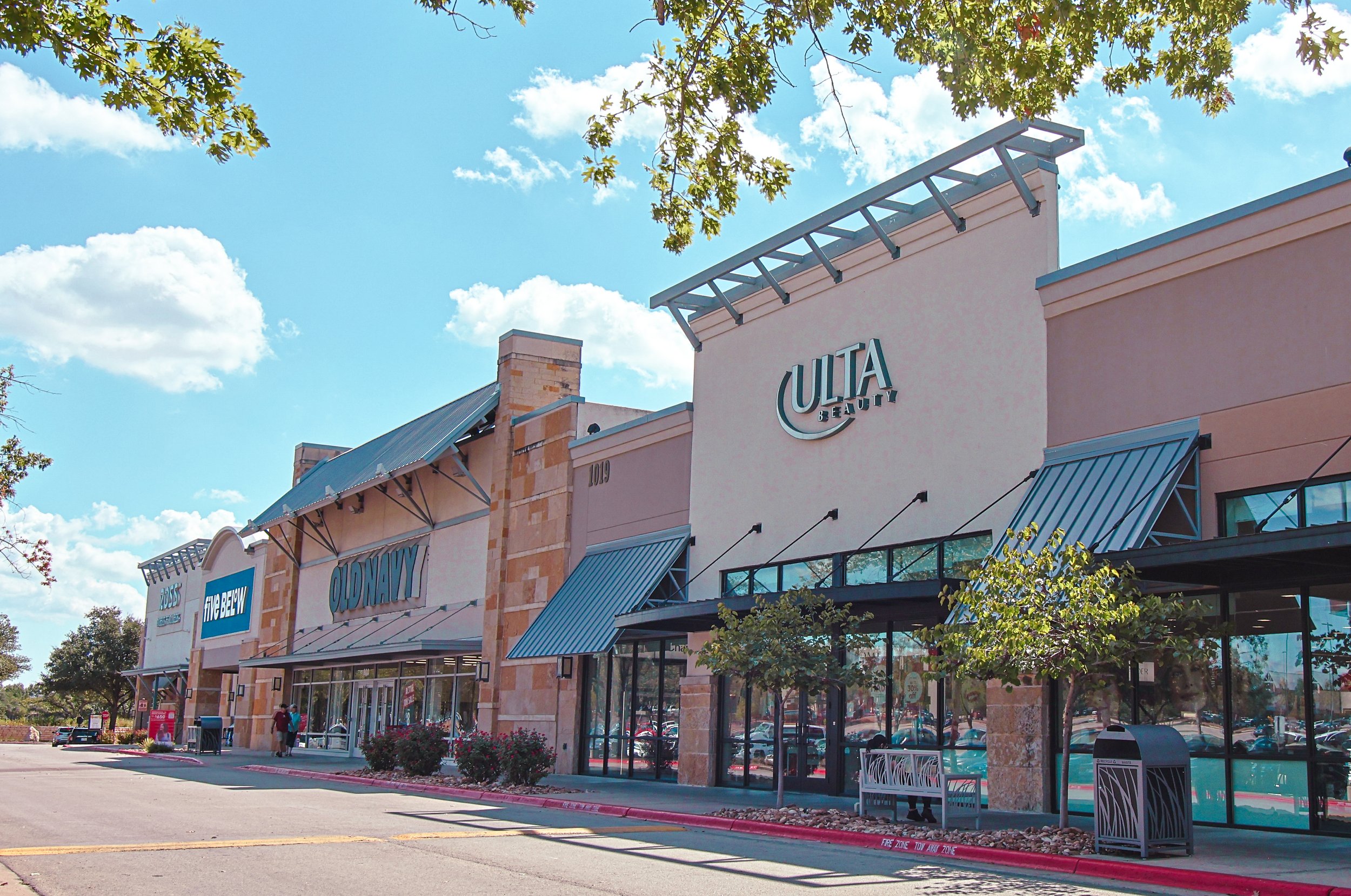 Wolf Ranch Town Center in Georgetown, TX, with stores Old Navy and Ulta Beauty, cars parked, a few people walking, trees, blue sky with clouds.
