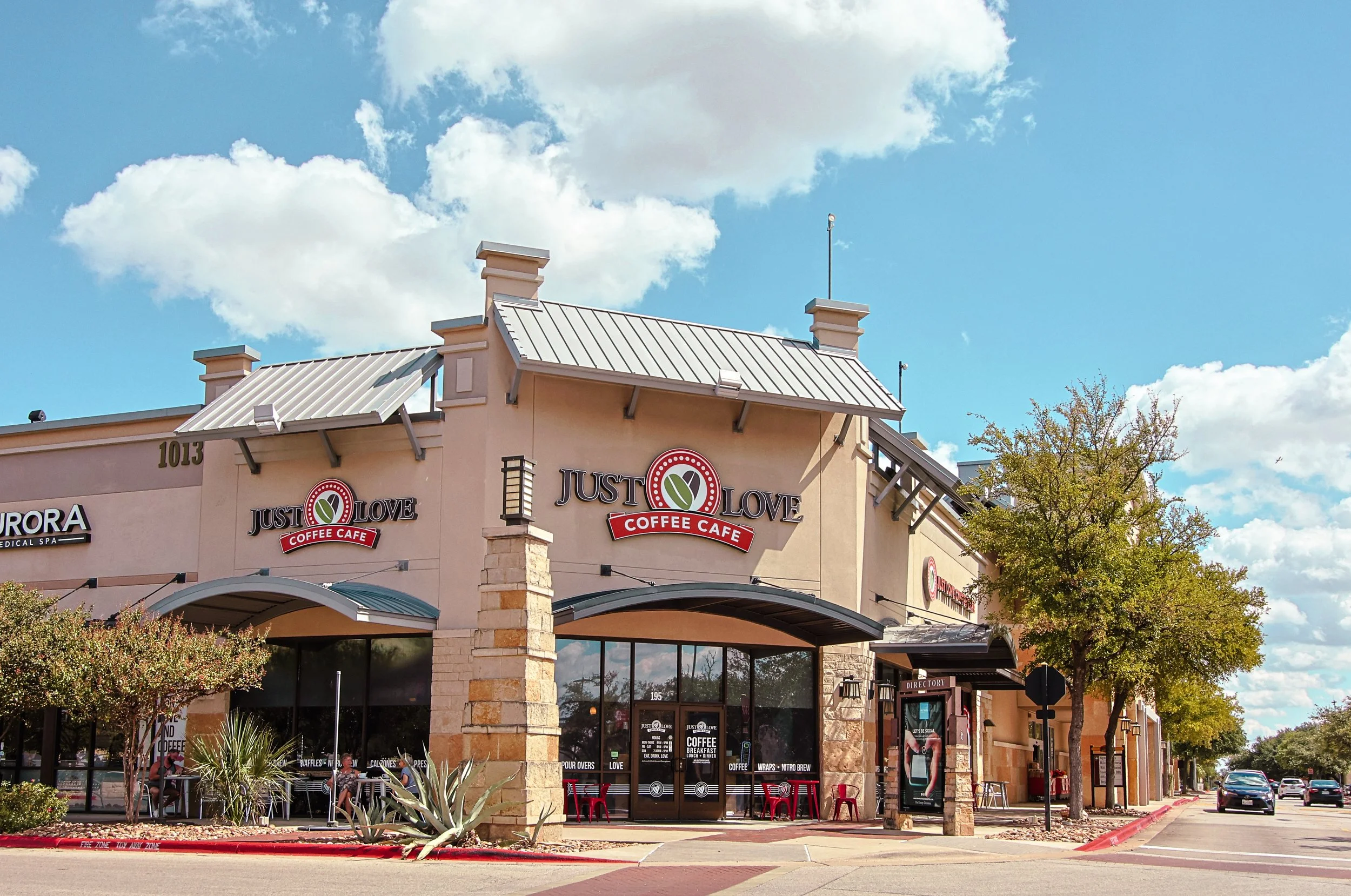 Exterior view of a Just Love Coffee Cafe coffee shop at Wolf Ranch Town Center in Georgetown, TX, with a sign displaying the cafe's name and logo, trees, parked cars, and a partly cloudy sky.