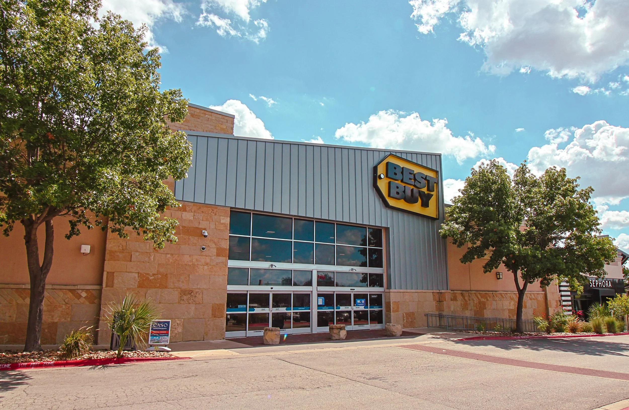 Exterior view of a Best Buy store at Wolf Ranch Town Center in Georgetown, TX, with large glass doors, trees on both sides, and a blue sky with clouds overhead.