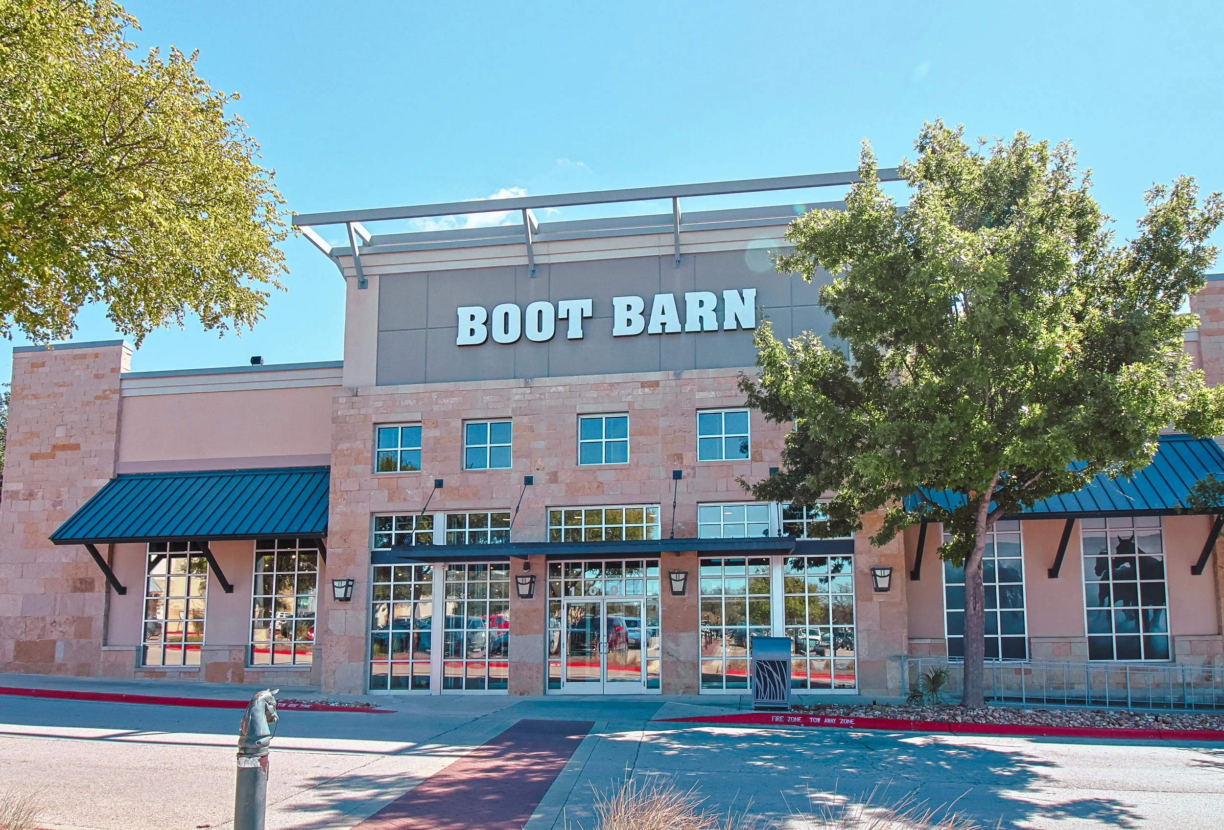 Front view of a building with a sign that reads "Boot Barn,"  at Wolf Ranch Town Center in Georgetown, TX, with large windows, and a tree in the foreground on a sunny day.