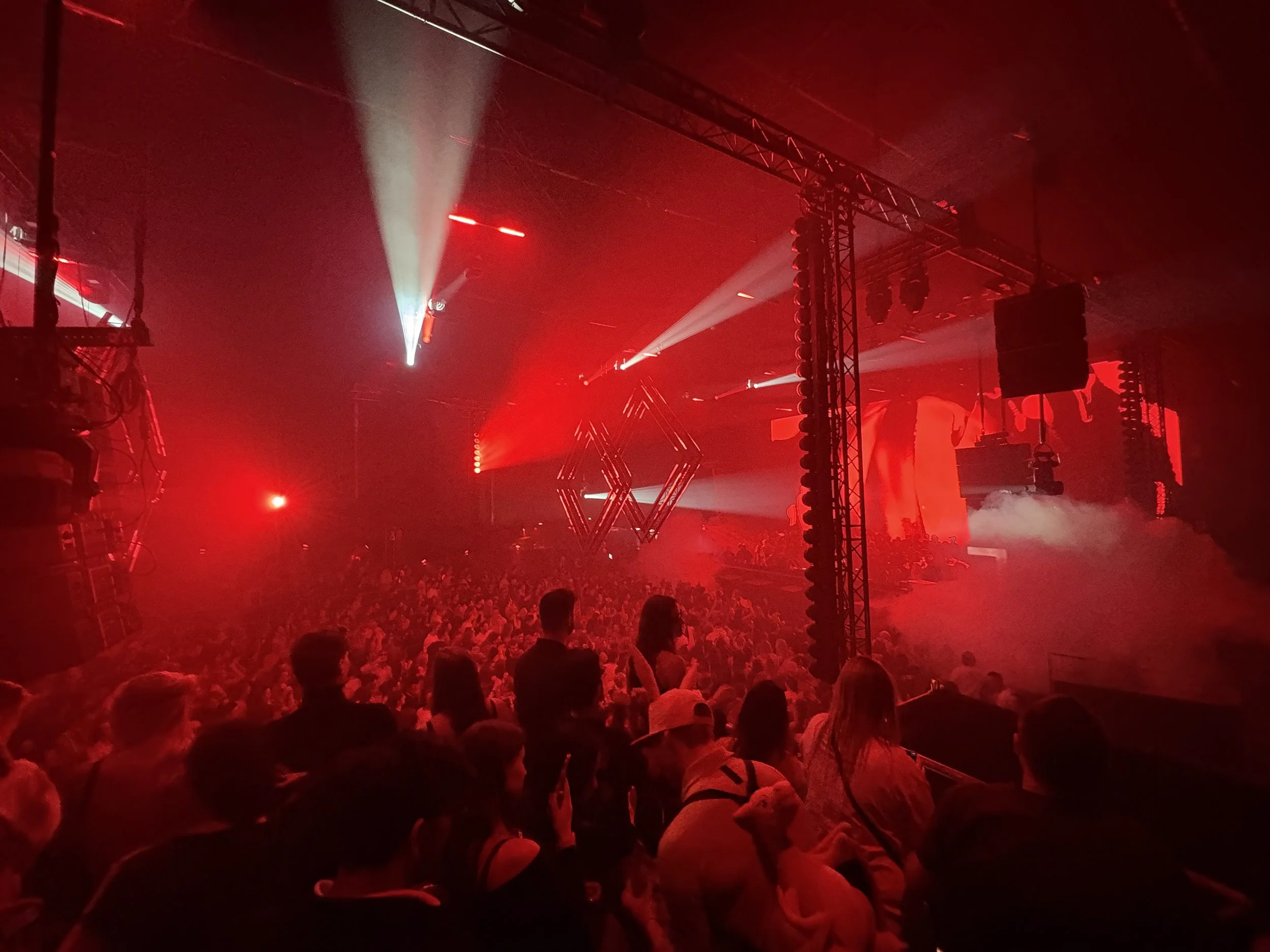 Crowd of people at a concert or nightclub with red and white stage lights, fog effects, and a large screen or display.