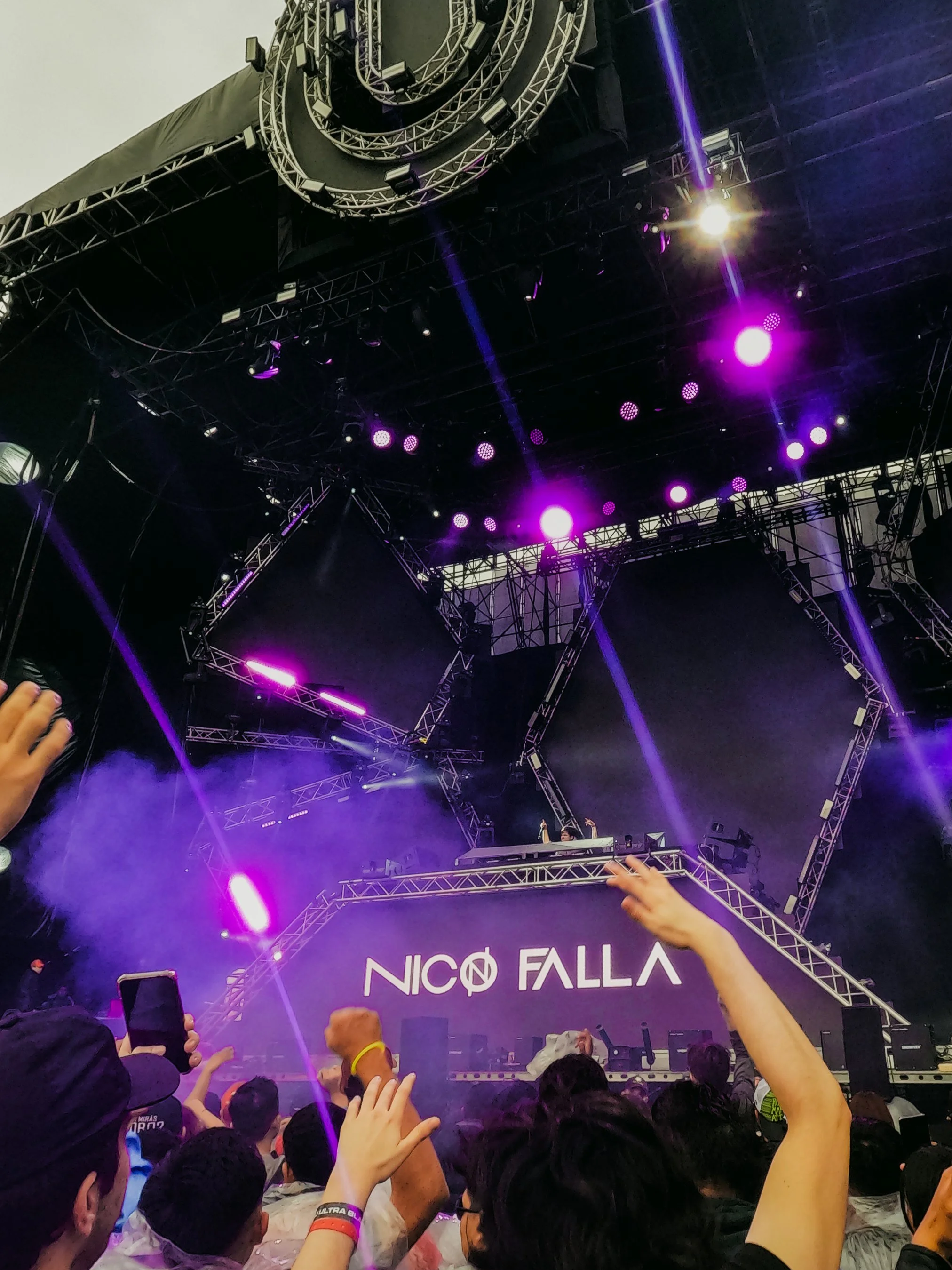 A concert scene with a stage illuminated by purple lights and LED screens displaying 'NICO FALLA.' The crowd is raising their hands, some holding smartphones, with visible smoke or fog effects.