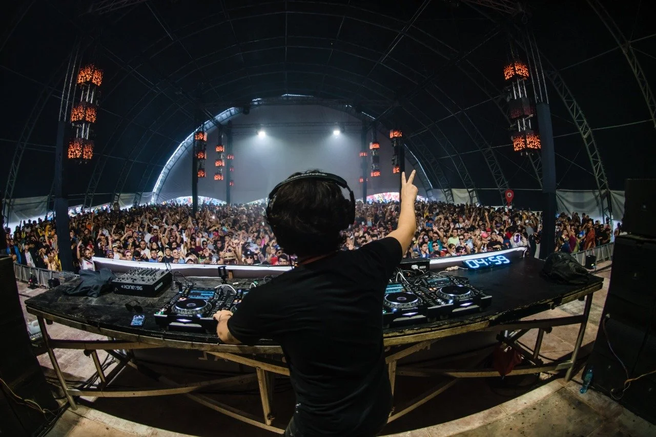 DJ performing at a large indoor music concert with a crowd of many fans, stage lights, and electronic equipment.