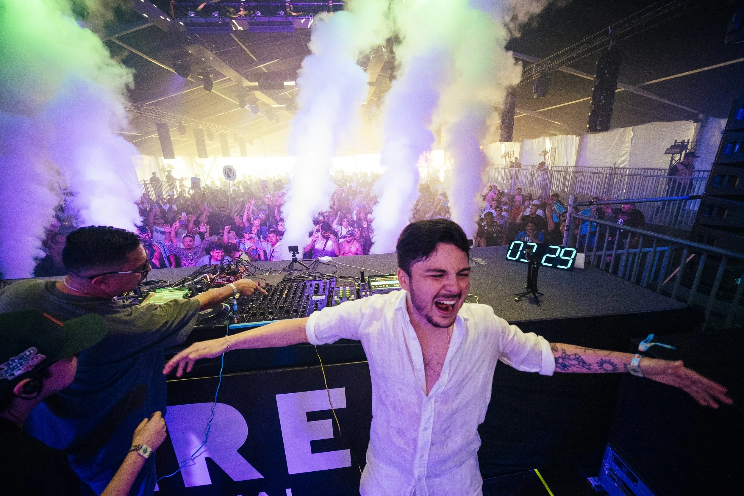 A DJ with a white shirt and tattooed arm dancing on stage at a crowded electronic music festival, with smoke effects and audience dancing in the background.
