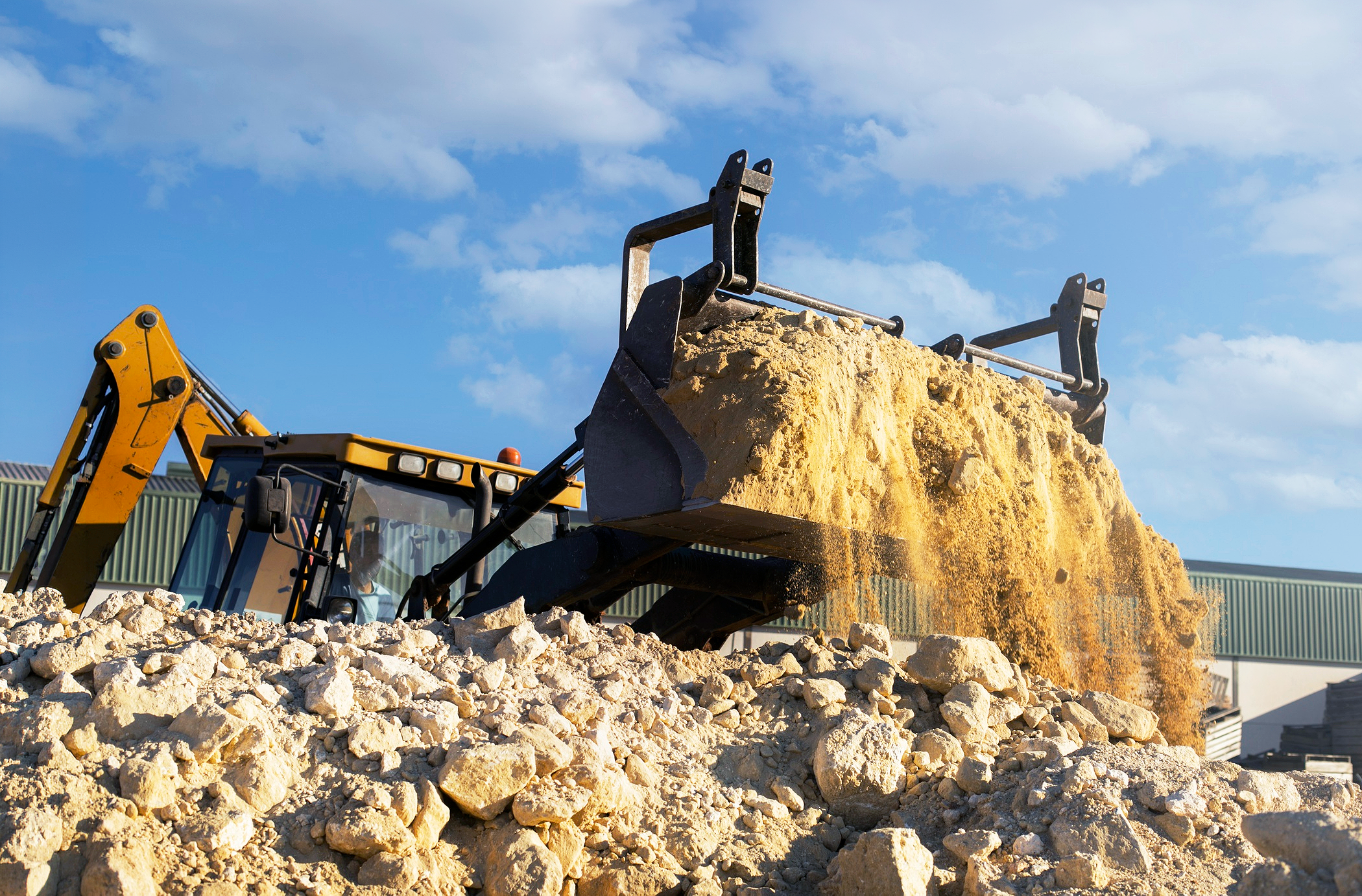 A construction excavator with a large scoop of dirt and rocks, lifting dirt with a bucket, against a blue sky with clouds.