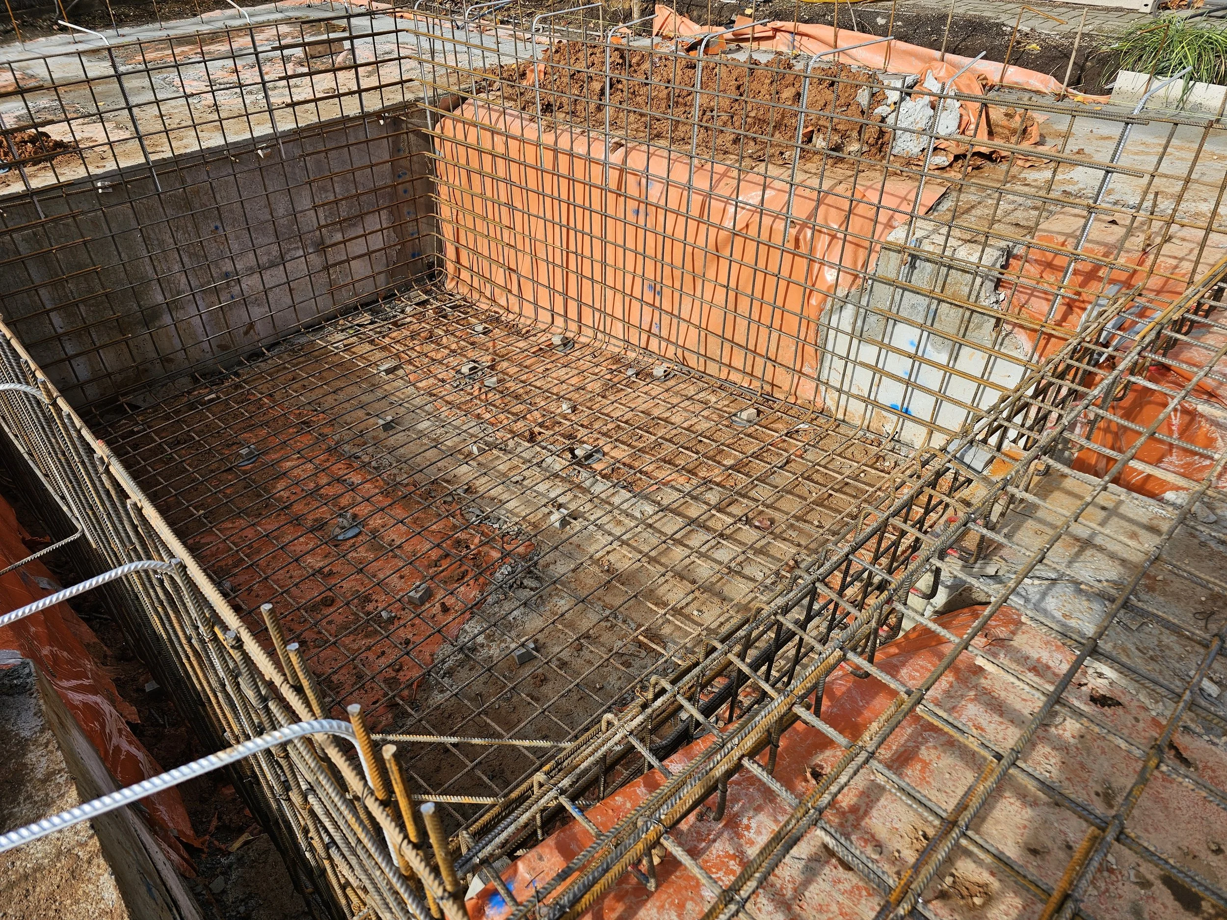 A construction site with concrete formwork and steel rebar for a foundation or footing, partially poured with concrete, surrounded by soil and construction materials.
