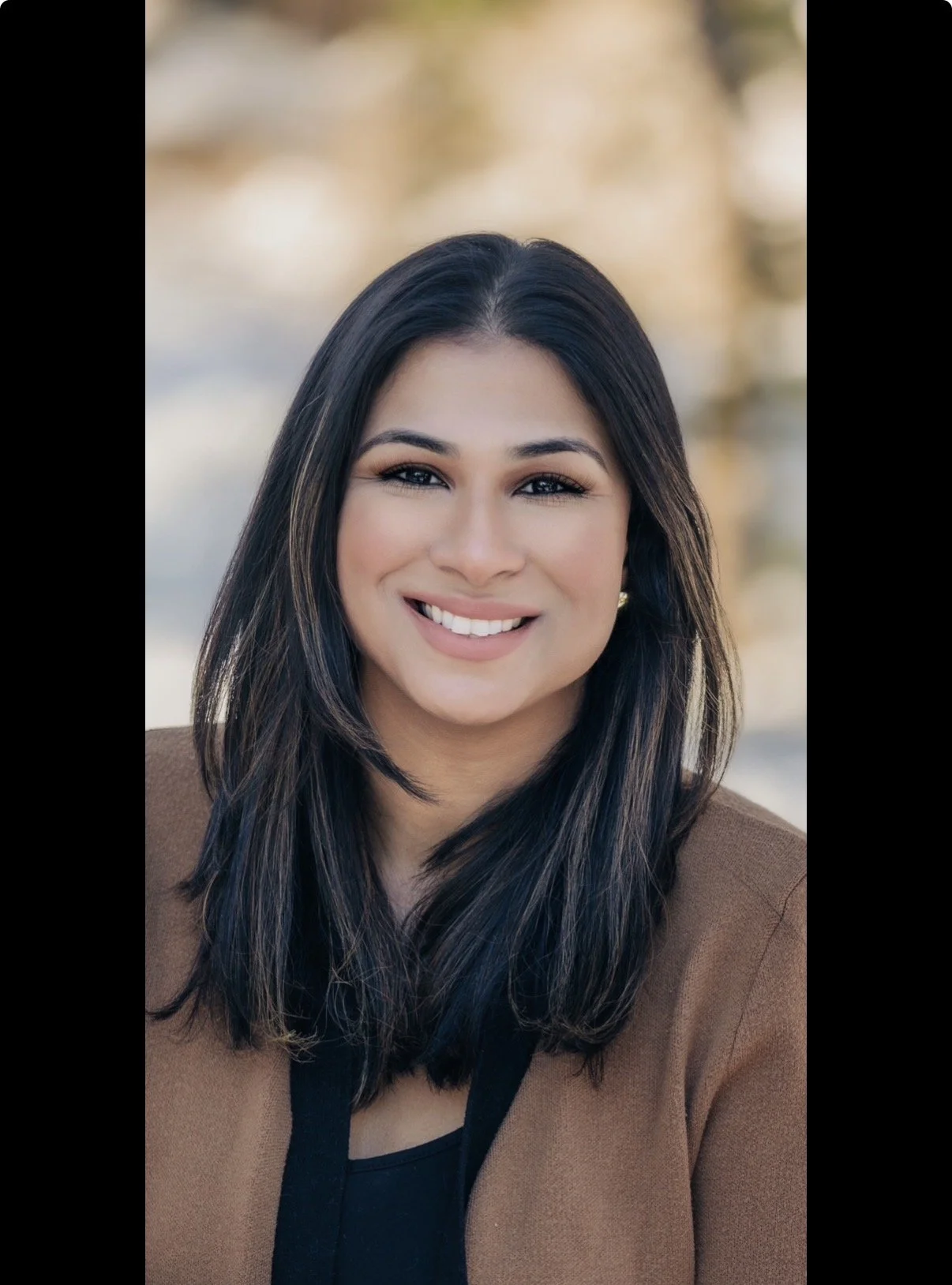 Portrait of a trauma therapist in Somerset, NJ  with dark hair, smiling, wearing a brown blazer and black top, with a blurred background.