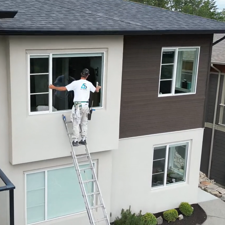A worker standing on a ladder installing or repairing a window on a two-story house with white and dark brown exterior siding.