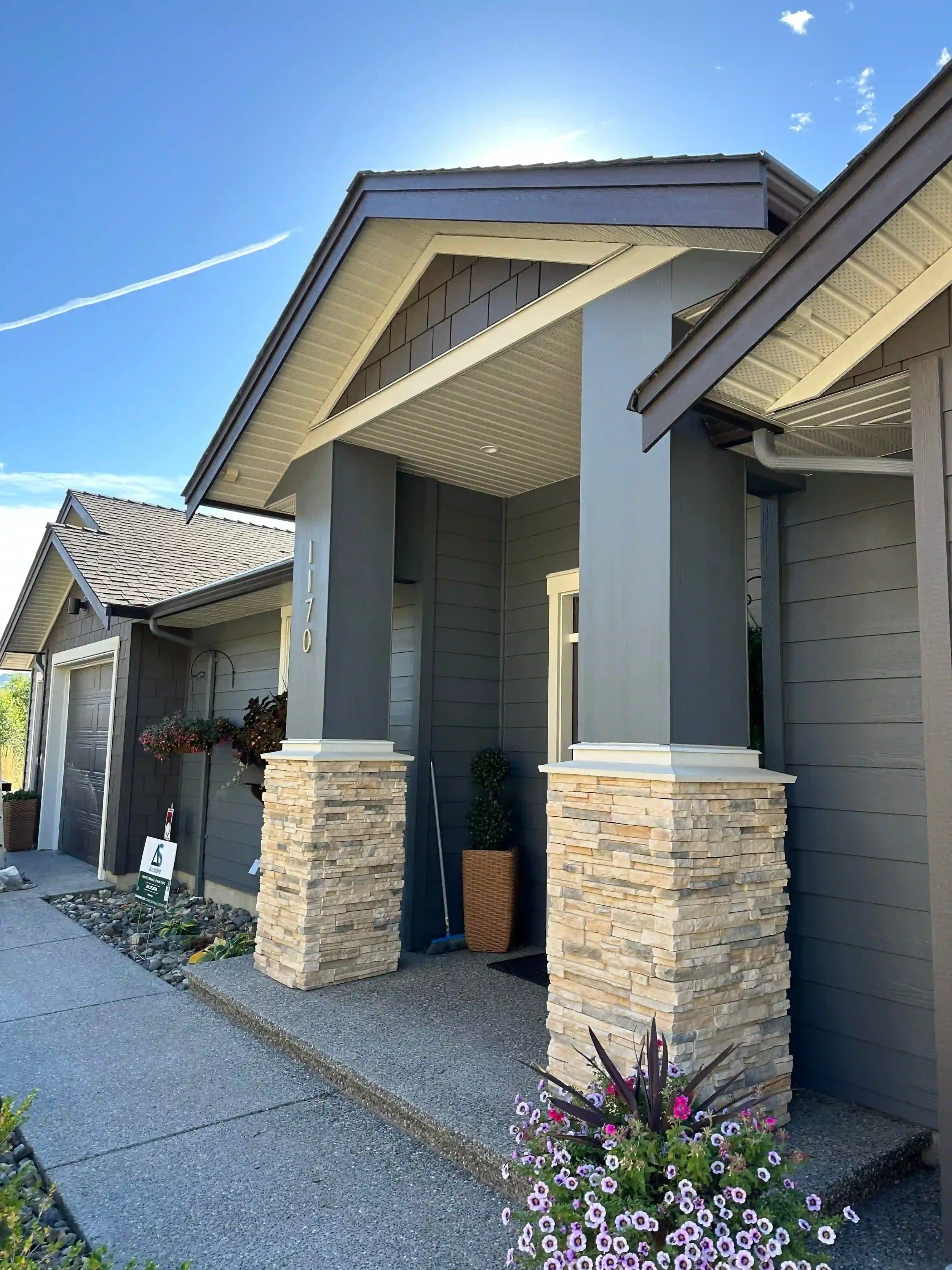 Front porch of a modern house with two stone pillars, gray siding, and a small potted plant, under a sunny blue sky.
