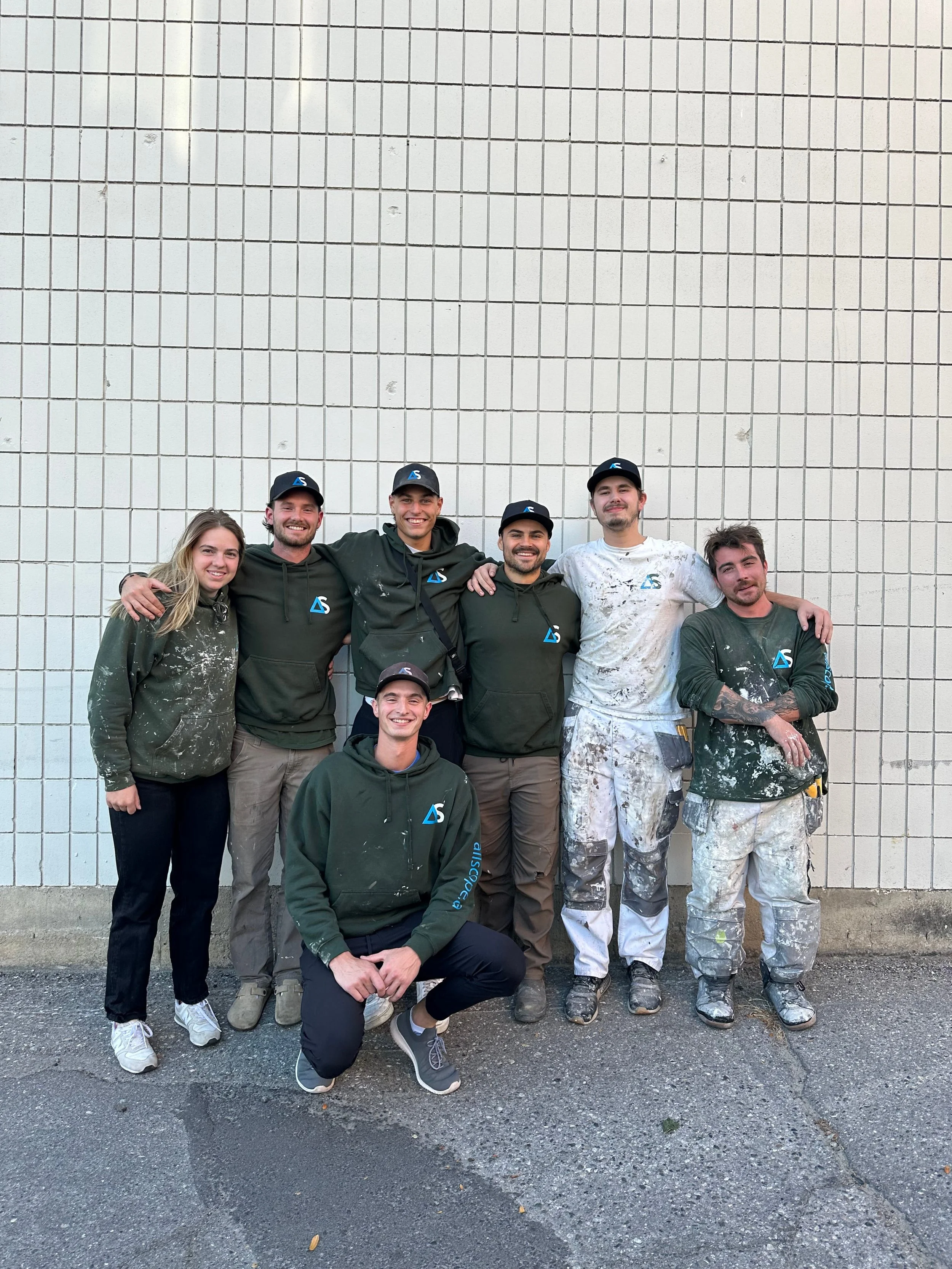 Group of seven young painters standing against a tiled wall, with some wearing paint-splattered work clothes and green hoodies, and others in dark hoodies with a logo, smiling and posing for the photo.
