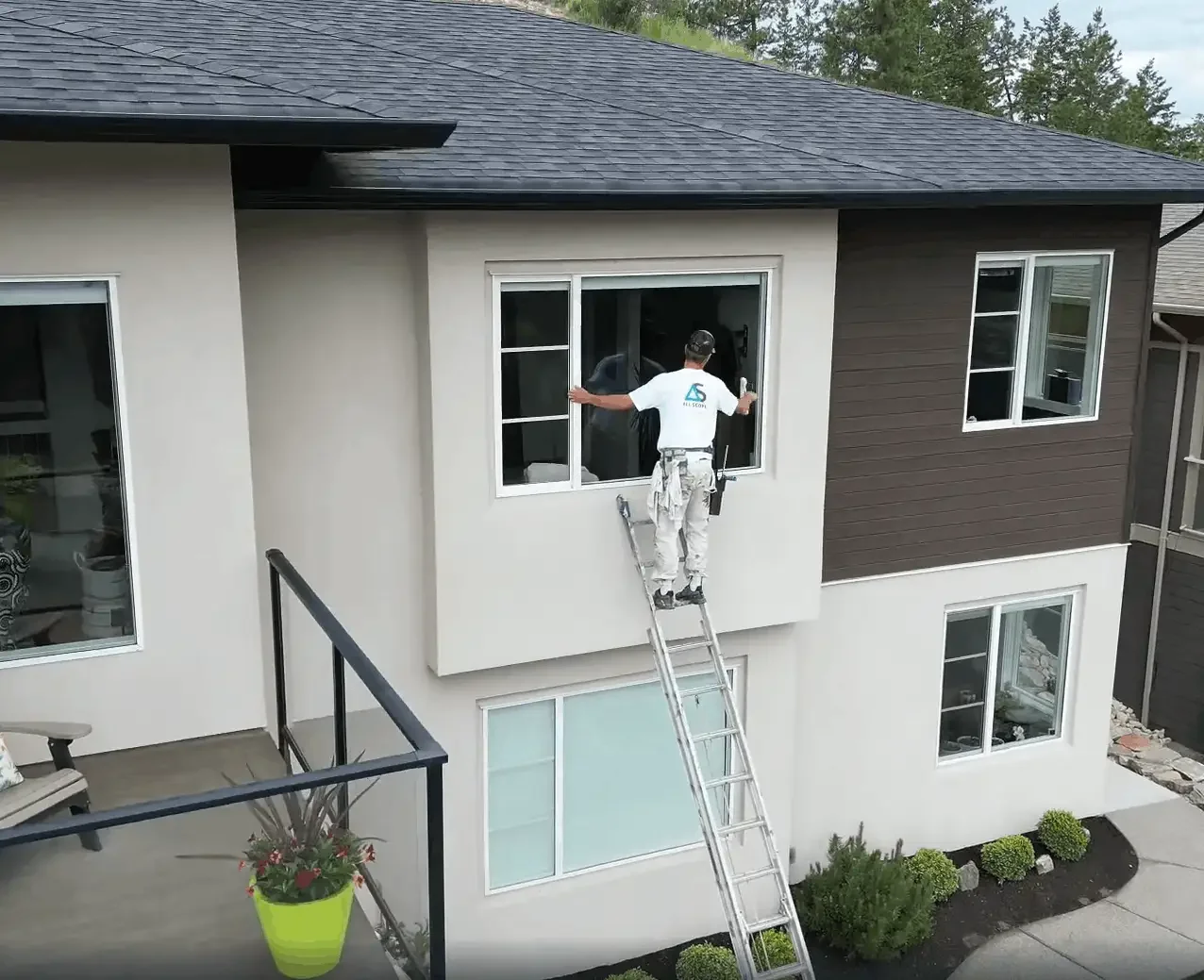 A worker standing on a ladder installing or repairing a window on a two-story house with white and dark brown exterior siding.