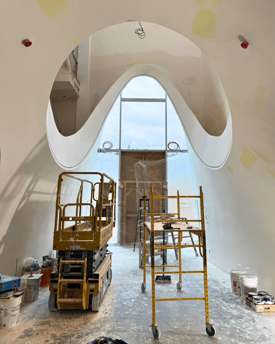 Under construction interior with a large abstract-shaped ceiling opening, construction lift, scaffolding, and building materials.