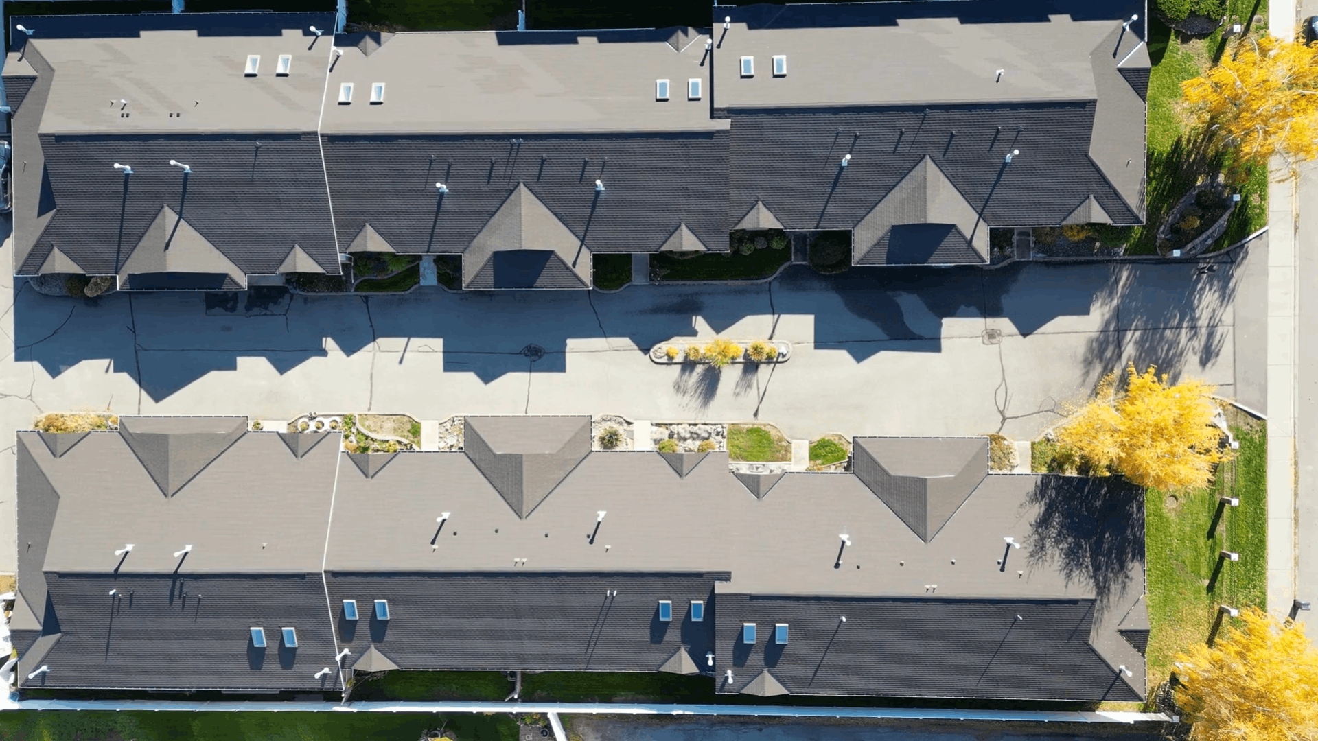 An aerial view of a residential apartment complex showing two rows of buildings with gray roofs, paved walkways, and landscaped areas with yellow-leaved trees.