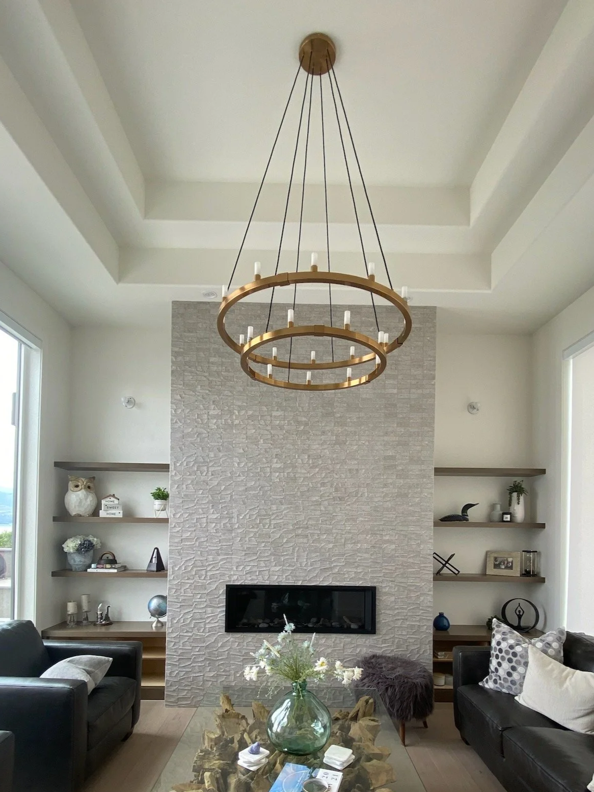 Modern living room with a white textured fireplace wall, black sofas, a glass coffee table with a green vase of white flowers, and built-in shelves on both sides of the fireplace with decorative items. A large gold circular chandelier hangs from the ceiling.