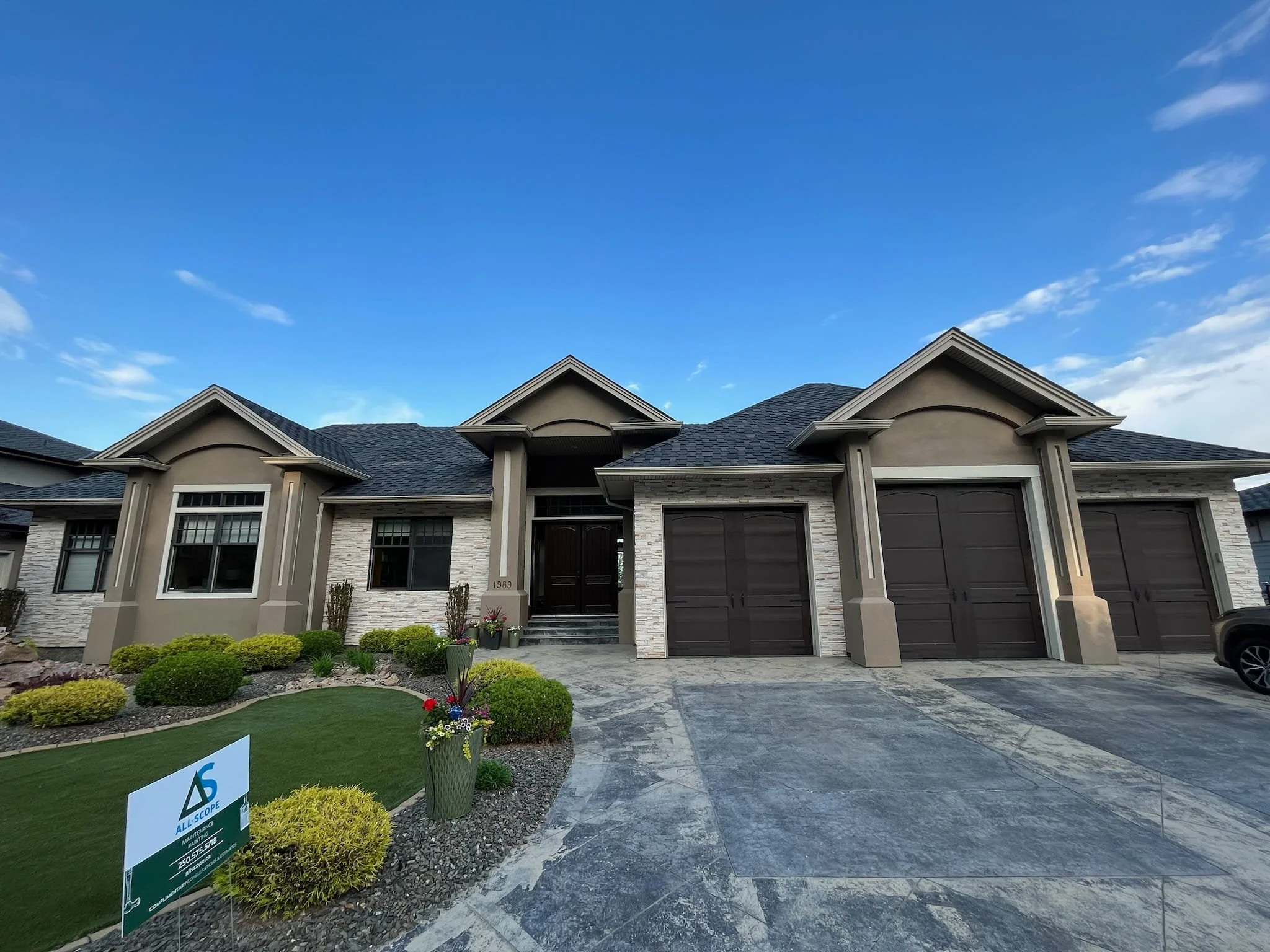 Modern house with three garage doors, landscaped front yard with bushes, flowers, and a lawn, under a blue sky with some clouds.